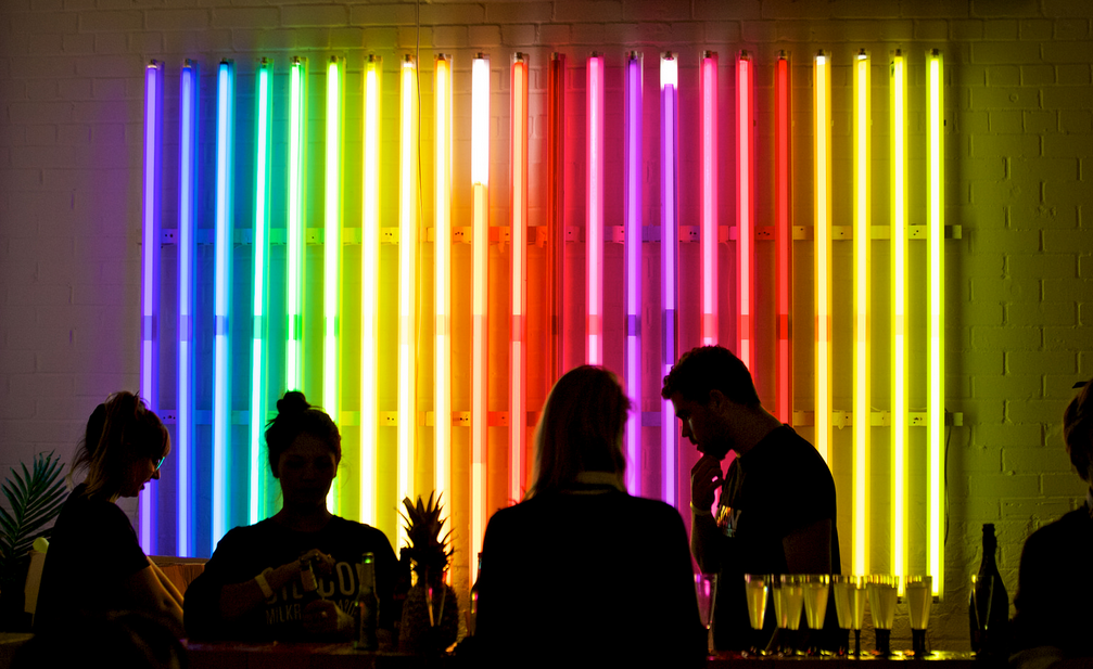 Group of people socializing in front of a colorful neon light art installation at night.