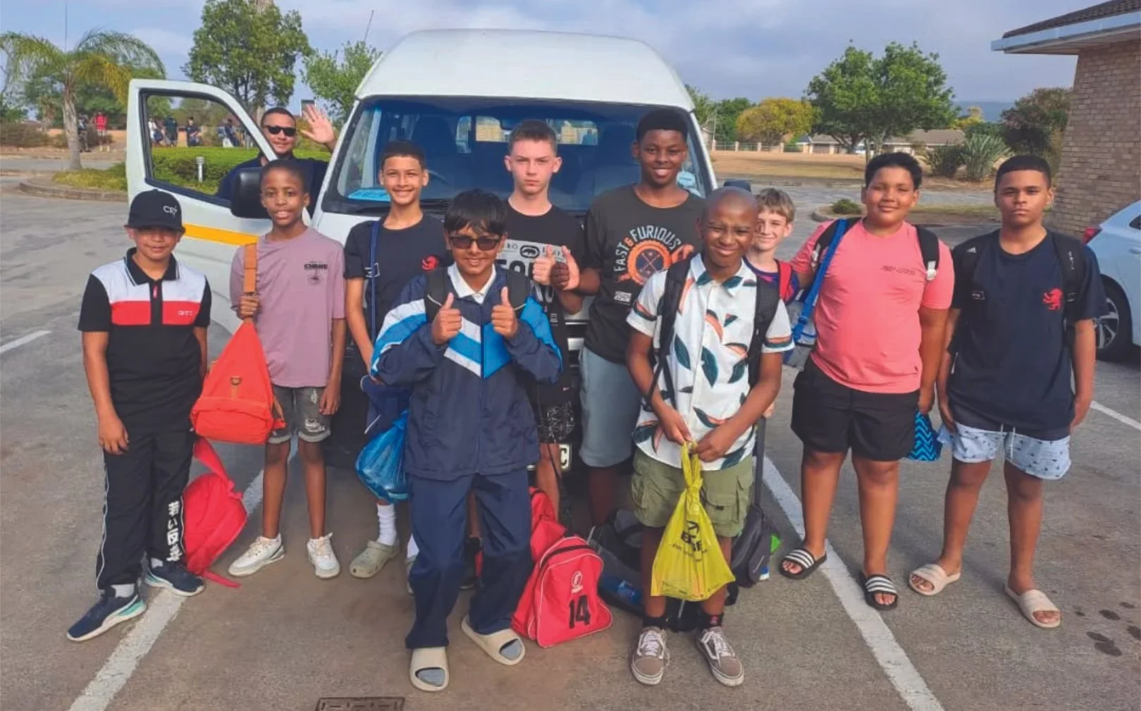 A group of children and one adult standing in a parking lot with backpacks, in front of a small van. The children are ready for a trip, and the adult is waving from the van.