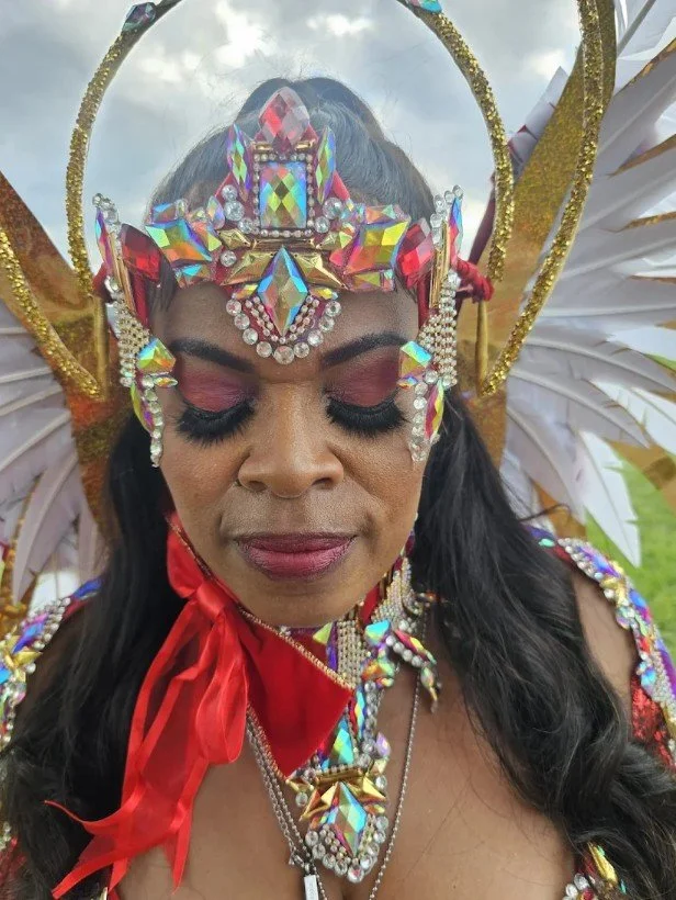 A woman with closed eyes wearing an elaborate costume with colorful gemstones, feathers, and gold accessories for a carnival or festival.