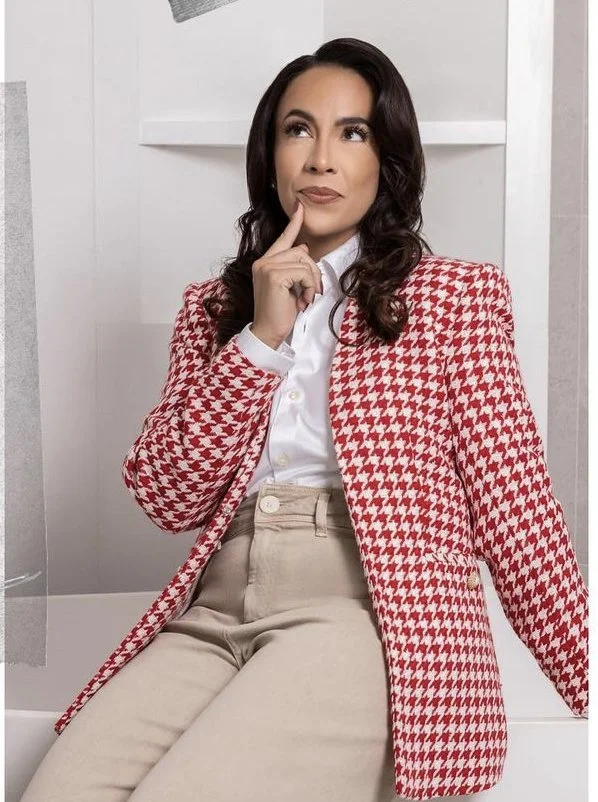 Woman with dark hair in a thoughtful pose, wearing a red and white houndstooth blazer, white shirt, and beige pants, sitting indoors.