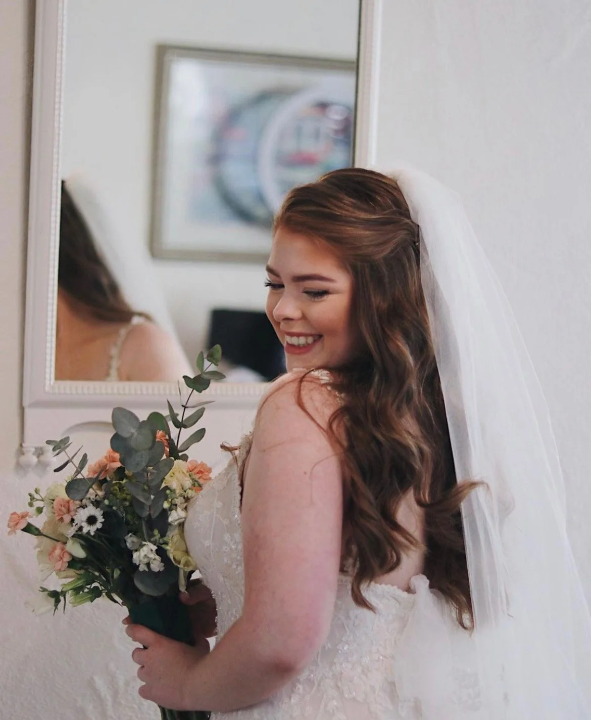 A smiling bride with long brown wavy hair wearing a wedding dress and veil, holding a bouquet of flowers, standing in front of a mirror.