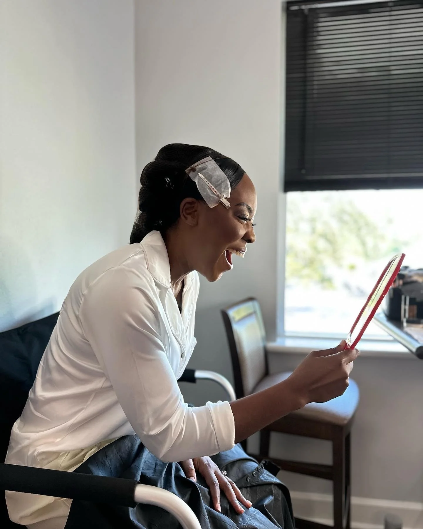 A woman with a bandage on her forehead is sitting in a chair, smiling and looking at a tablet she is holding.