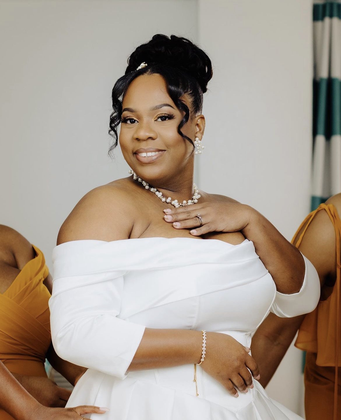 A woman dressed in a white off-shoulder gown, elegant jewelry, and styled black hair, smiling at the camera.