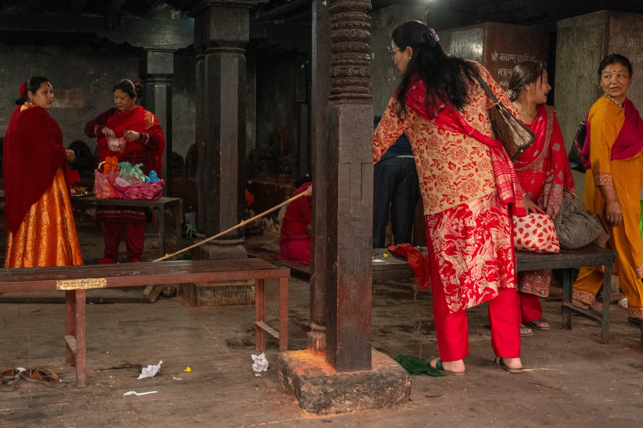 A group of women in colorful traditional Indian sarees and dresses standing and talking inside a dark, rustic temple or shrine with wooden pillars and stone floor.