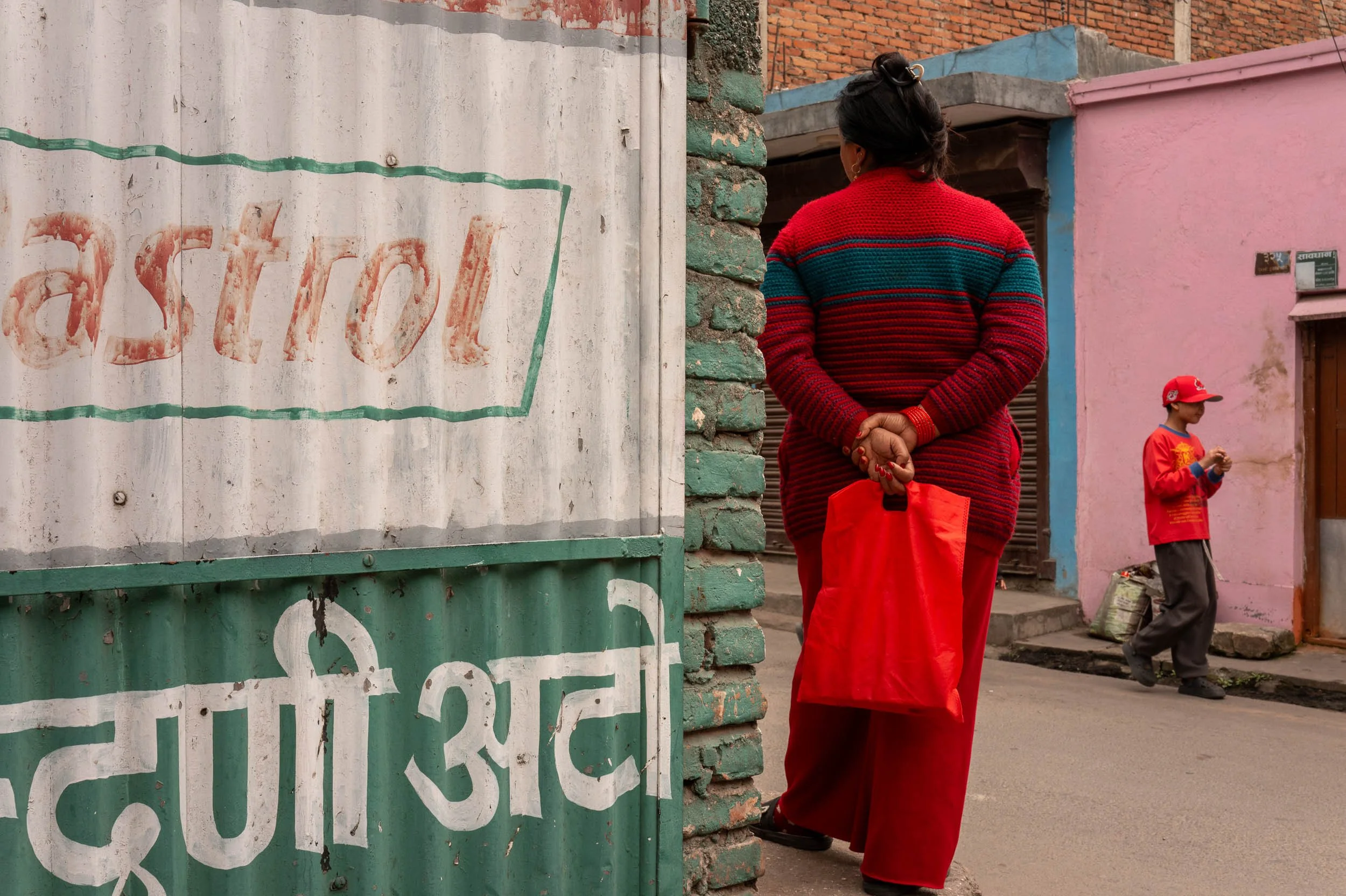 A woman dressed in a red sweater and red skirt standing on a street, with her hands clasped behind her back, facing away from the camera. She is near a painted wall and a brick building, with a young boy in red clothing and a cap standing nearby.