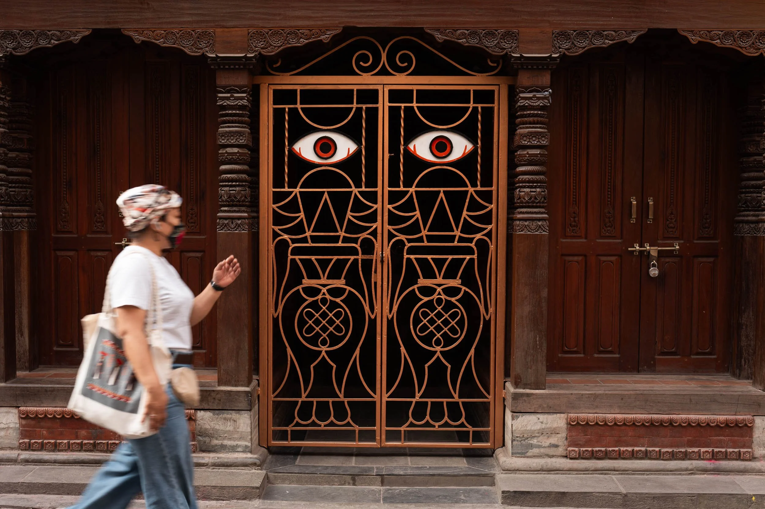Wooden door with intricate carved details and a decorative metal gate featuring eye-shaped designs, with a woman walking past and wearing a face mask and headscarf.