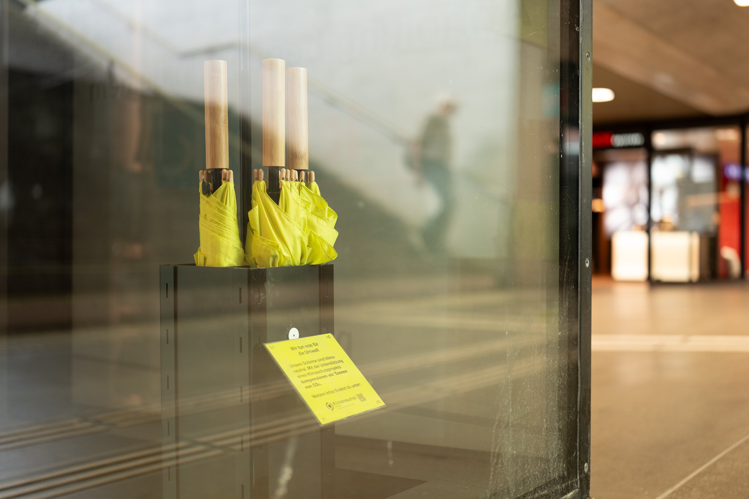 A row of three yellow umbrellas leaned against the glass wall of an elevator or enclosed space, with blurred indoor background.