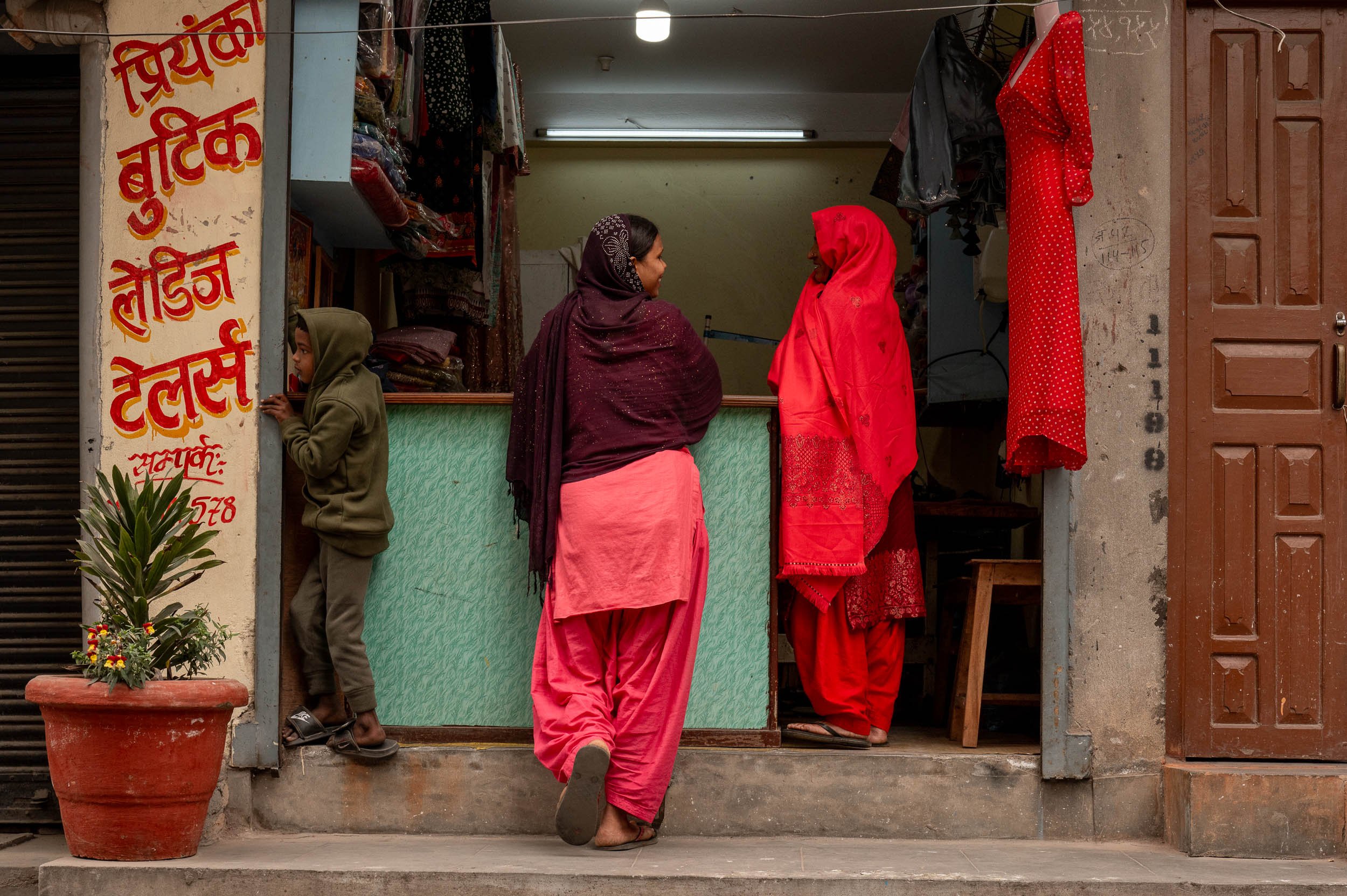 Two women and a child standing outside a shop in an urban setting. The women are wearing traditional Indian clothing and headscarves, and they are engaged in conversation. The child is wearing a hoodie and standing near the shop entrance. The shop has colorful fabric or clothing items on display, a potted plant with flowers, and a sign written in Hindi on the left side of the image.