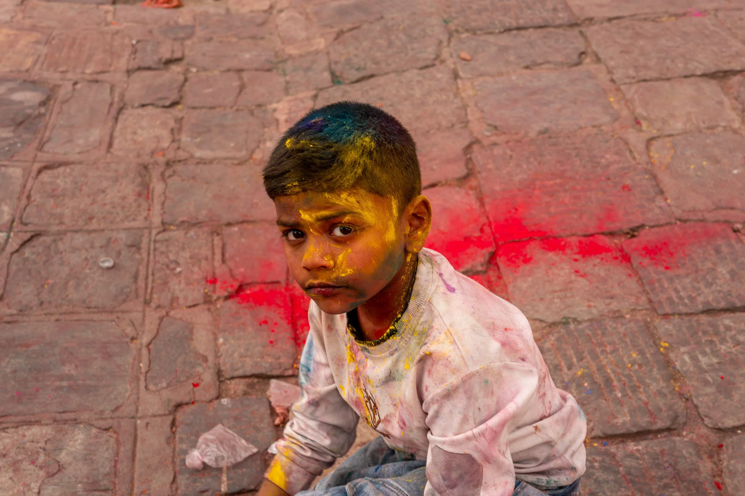A young boy with colored powder on his face and clothes, looking up at the camera, sitting on a brick pavement during a celebration involving colorful powders.