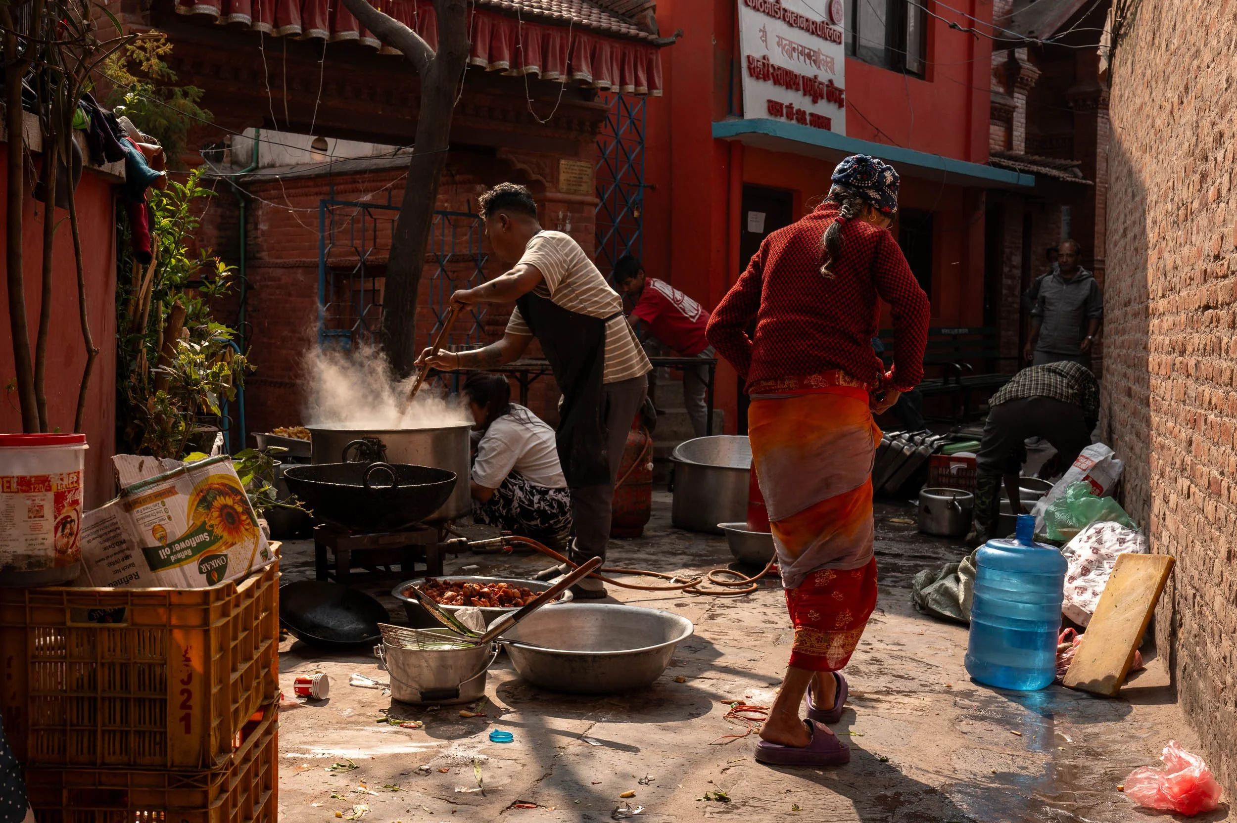 Street food stall in Nepal with women cooking over an open fire, large cooking pots, and various supplies around on a narrow alley.