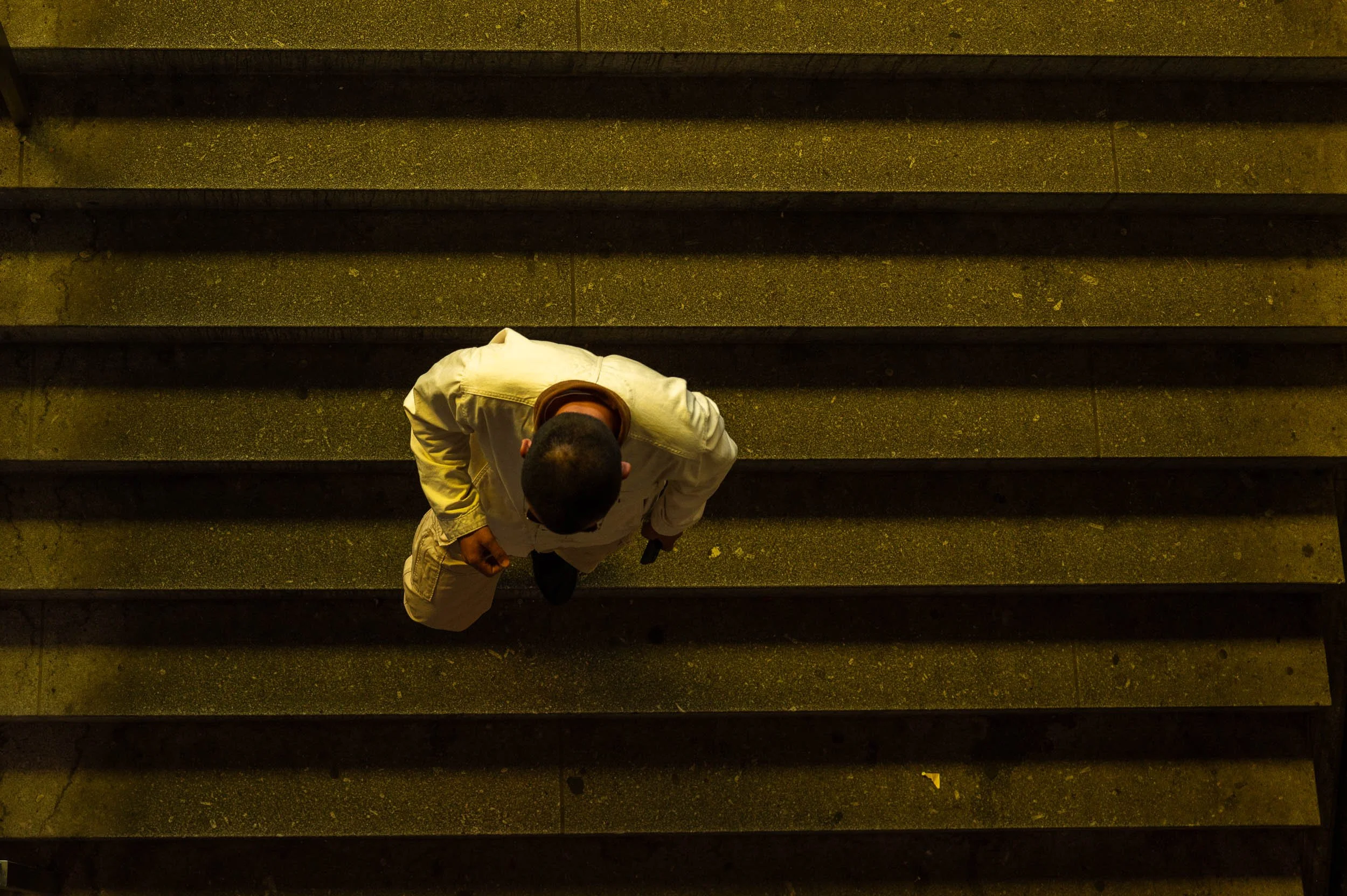 A person wearing a beige jacket, with short dark hair, viewed from above, walking up a set of concrete stairs at night, holding a phone in their left hand.