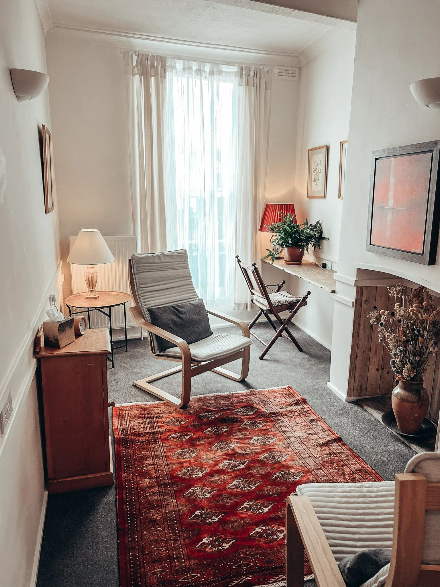 A cozy living room with natural light streaming through a large window covered with sheer white curtains. The room includes a white armchair with a black cushion, a small wooden side table with a table lamp, a vintage patterned red rug, some framed artwork on the walls, and a potted plant on a ledge next to the window. There is a mixture of modern and rustic decor, with a small wooden cabinet and a fireplace mantel.