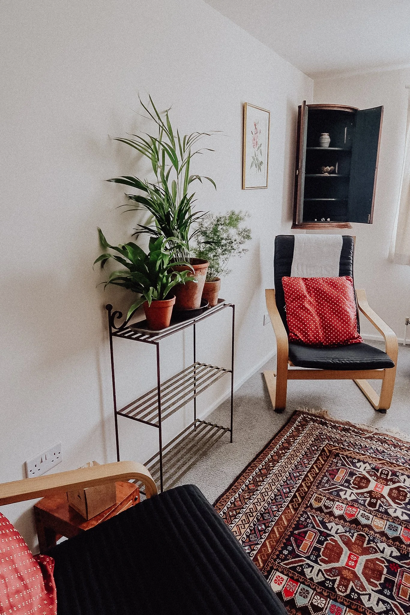 A cozy living room corner with a black armchair, red patterned pillow, wooden side table, potted plants on a black metal stand, a framed floral painting, a wooden cabinet, a colorful patterned rug, and beige carpet.