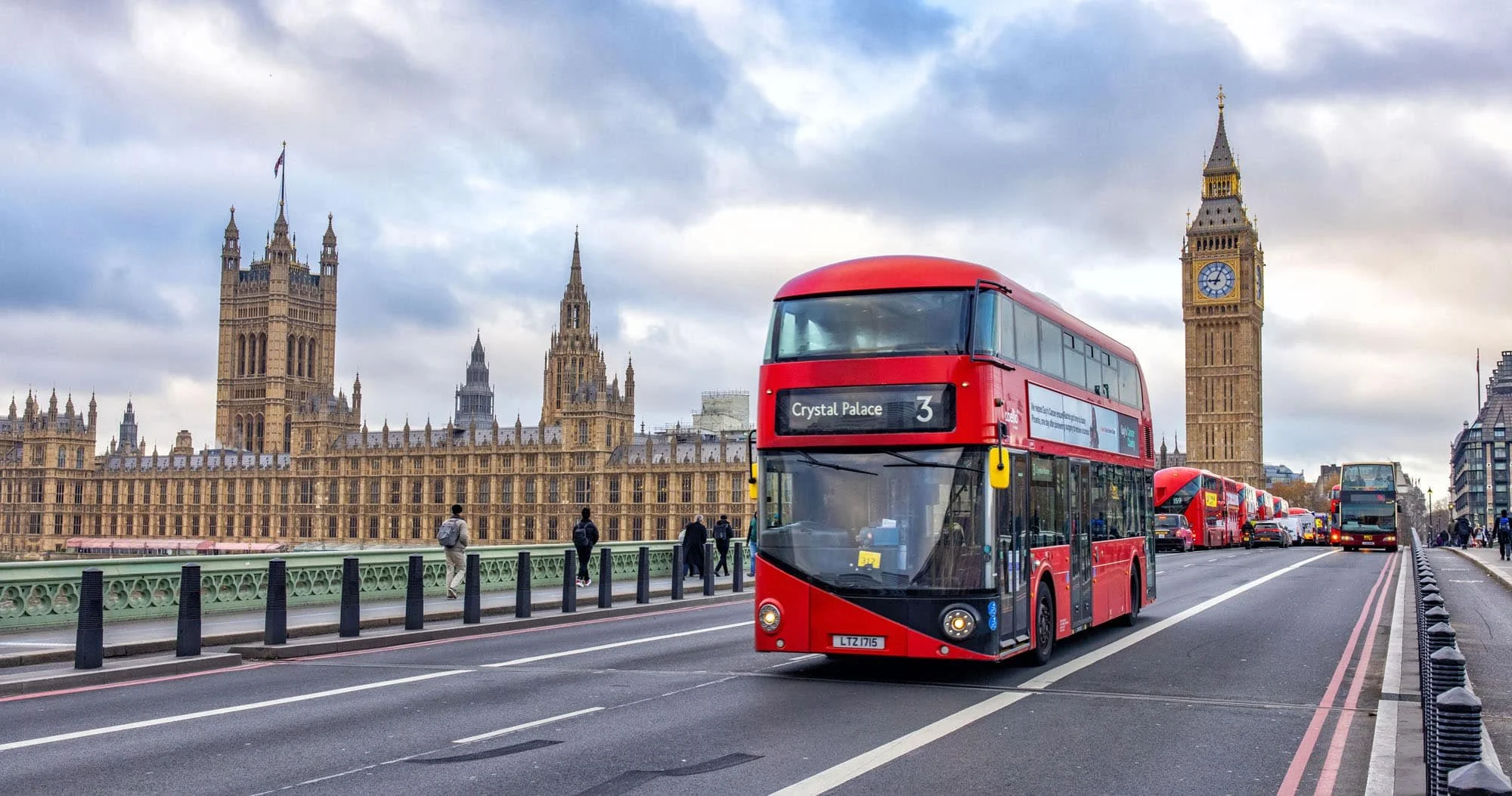 Red double-decker bus on a bridge with Big Ben and the Houses of Parliament in London in the background, cloudy sky, pedestrians walking on the bridge, and additional buses in the lane.