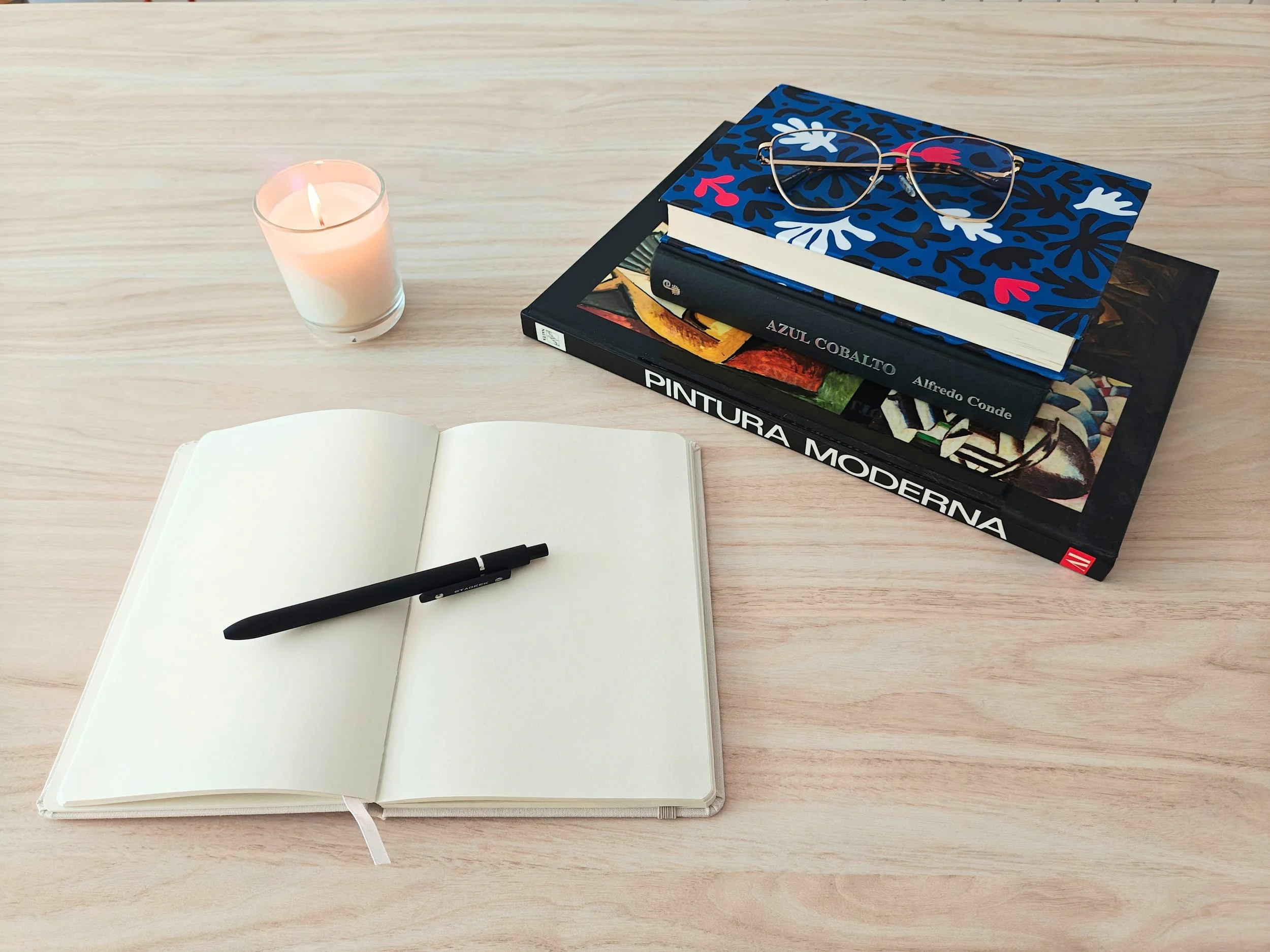 Open blank notebook with a black pen on a wooden desk. To the right, there is a stack of art books, topped with a pair of gold wire-framed glasses. The items are accompanied by a lit candle to the left.