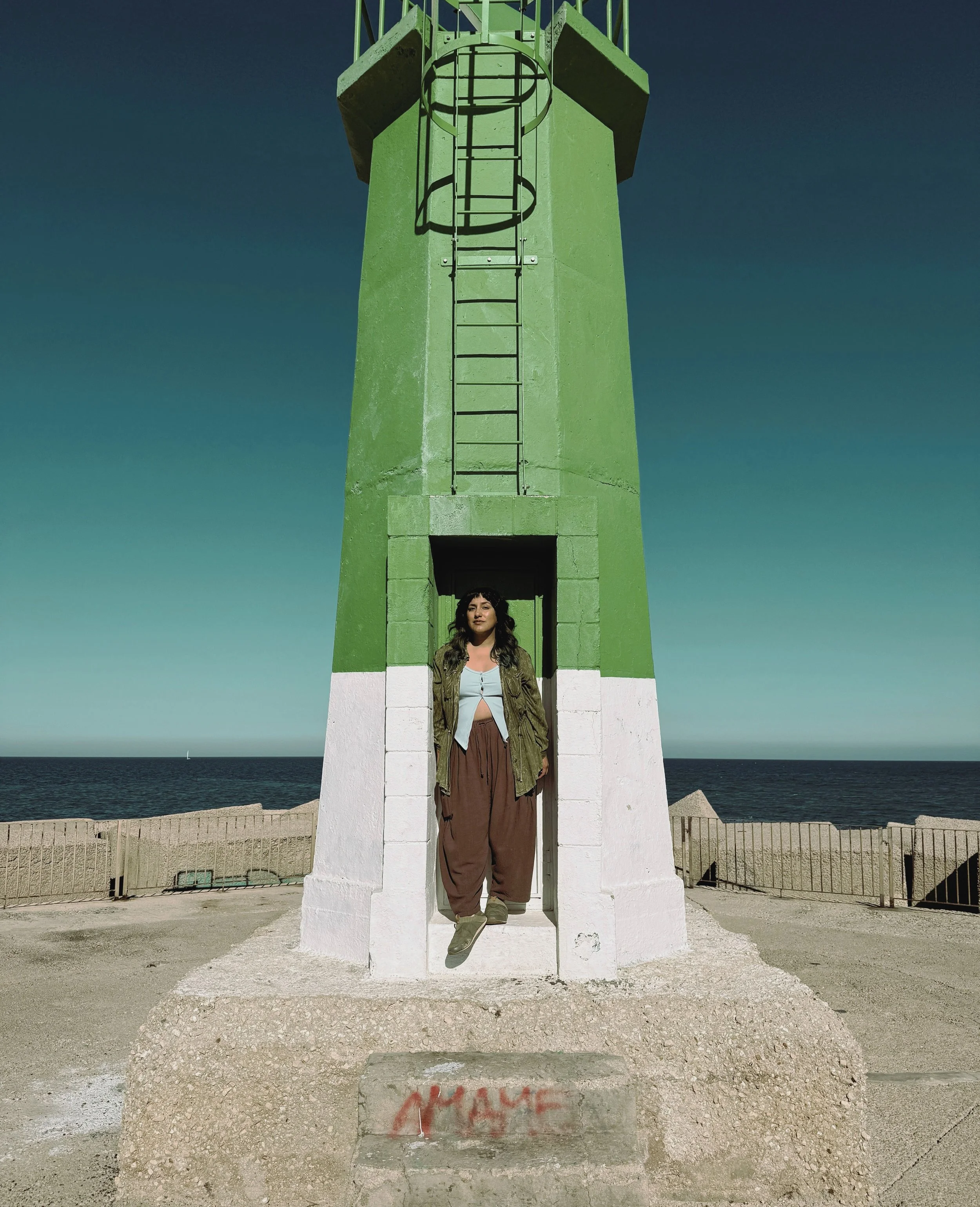 A woman standing at the base of a green and white lighthouse with a ladder attached to it. The backdrop shows an ocean with a sailboat in the distance. The woman is wearing a green jacket and loose brown pants, looking at the camera.