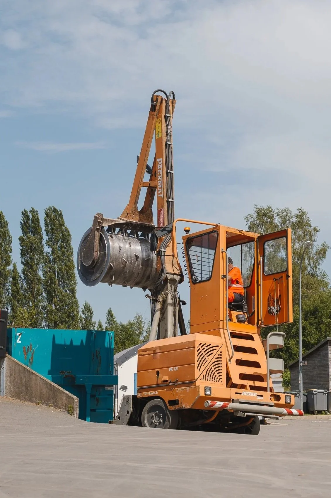 Une machine de construction orange avec un conducteur en tenue de sécurité orange, utilisant une excavatrice pour des travaux en extérieur. Arbres et ciel dégagé en arrière-plan.