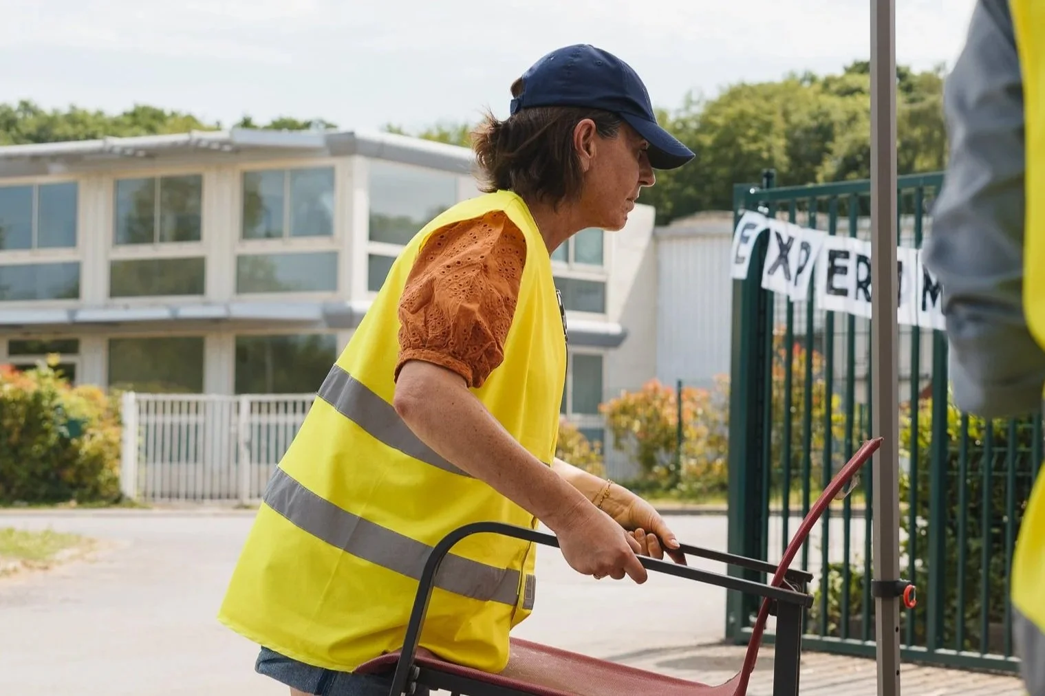 Une femme portant un gilet jaune réfléchissant déplace une chaise de bain sur un trottoir, en extérieur, près d'une clôture avec un bâtiment moderne en arrière-plan.