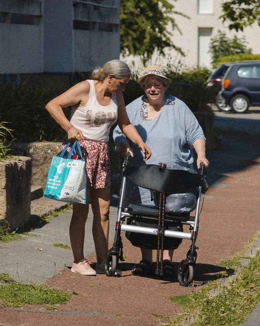 Deux femmes âgées, l'une aidant l'autre avec une déambulateur, marchant sur un trottoir en extérieur.