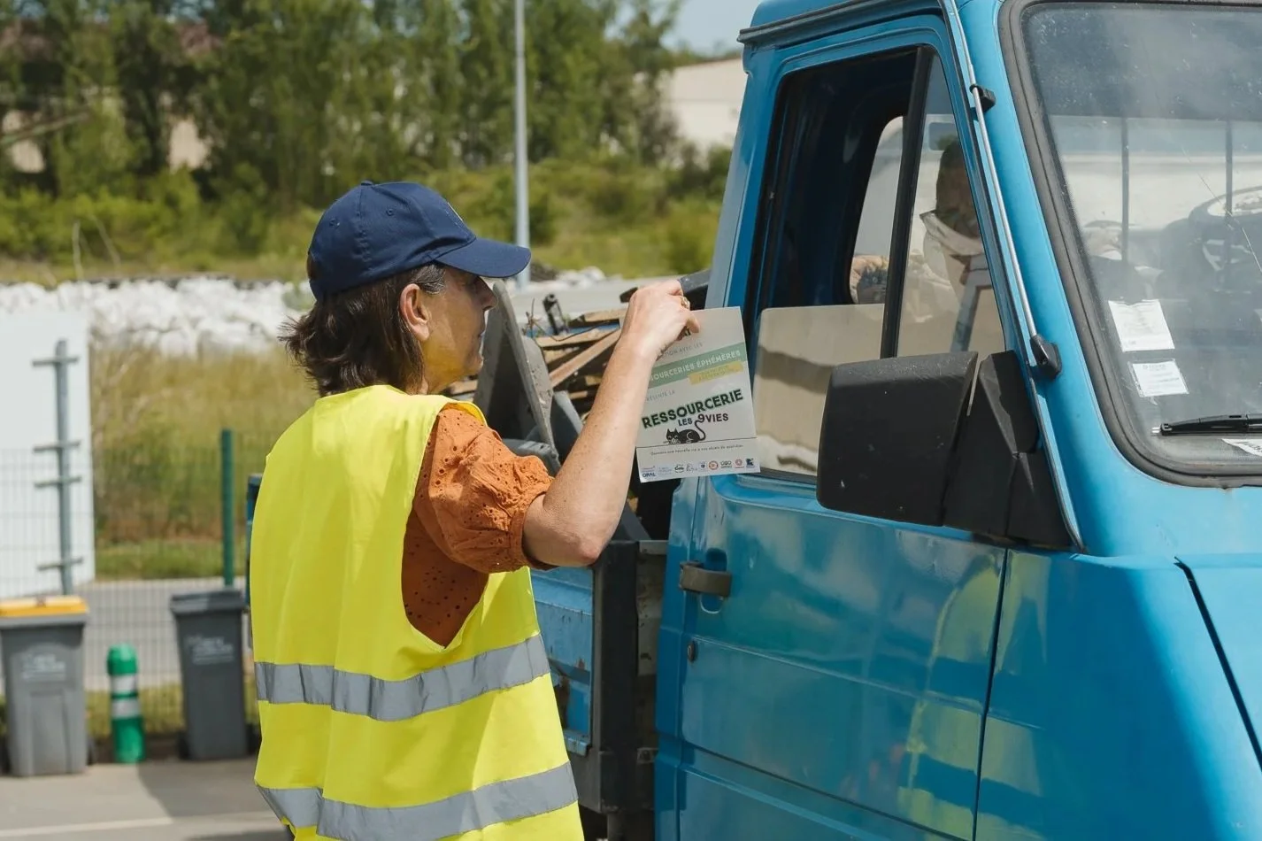 Une femme avec un gilet jaune distribue un flyer dans une camionnette bleue.