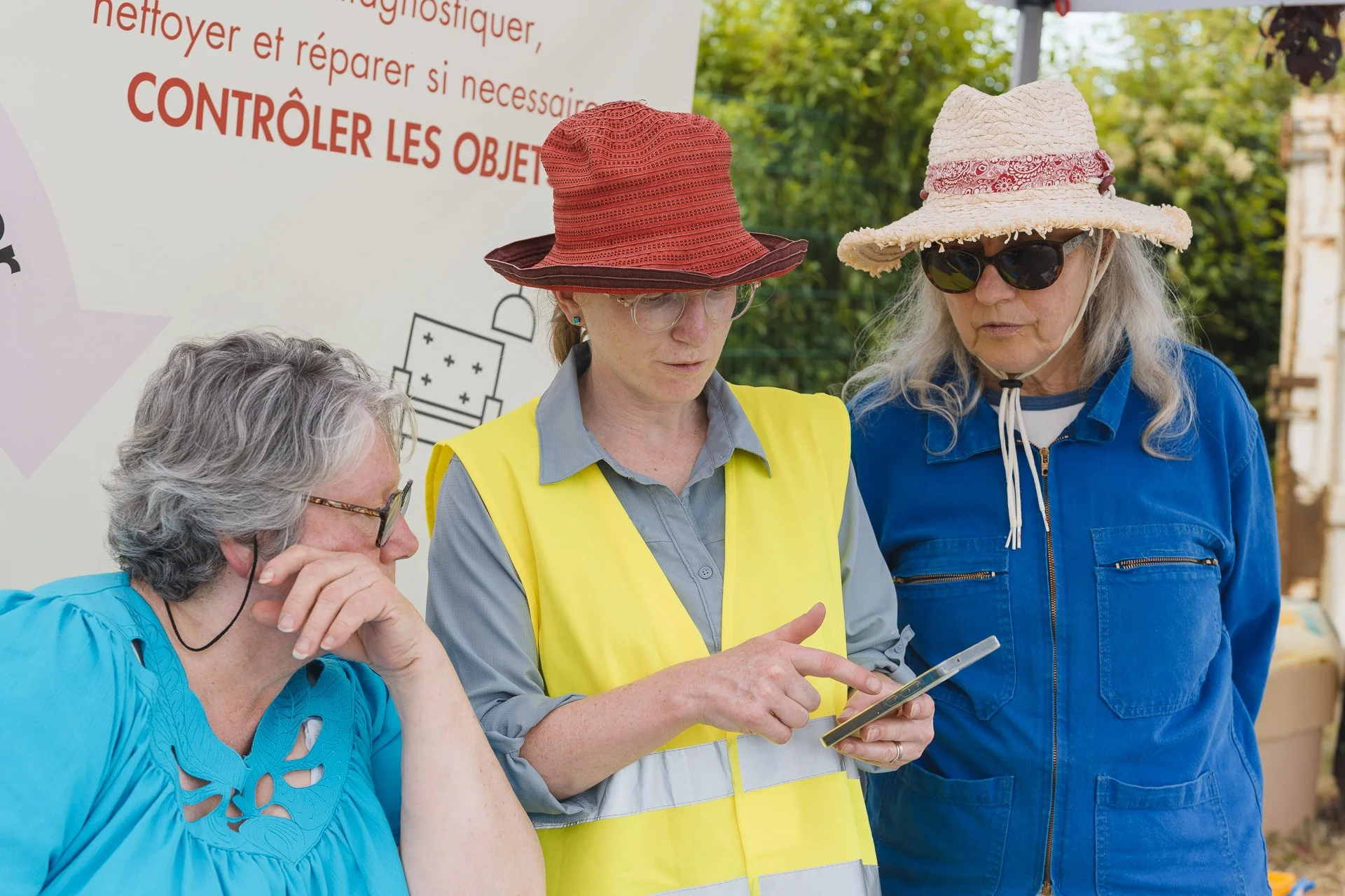 Trois femmes portant des chapeaux et lunettes de soleil discutant autour d'une tablette, avec un panneau informatif en arrière-plan.