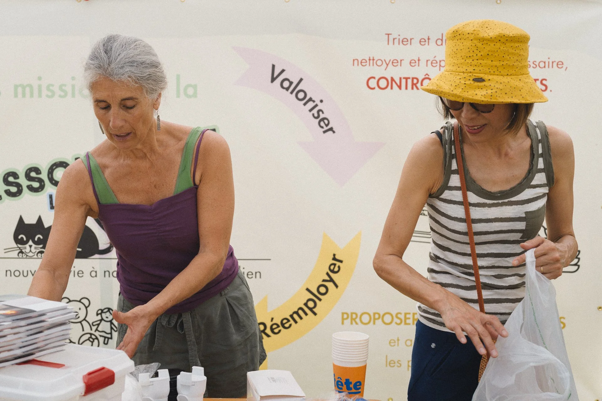 Deux femmes âgées, l'une avec des cheveux gris portant un haut violet et l'autre avec un chapeau jaune et des lunettes, participent à une activité ensemble, probablement dans un centre communautaire ou un événement public. Elles manipulent des objets