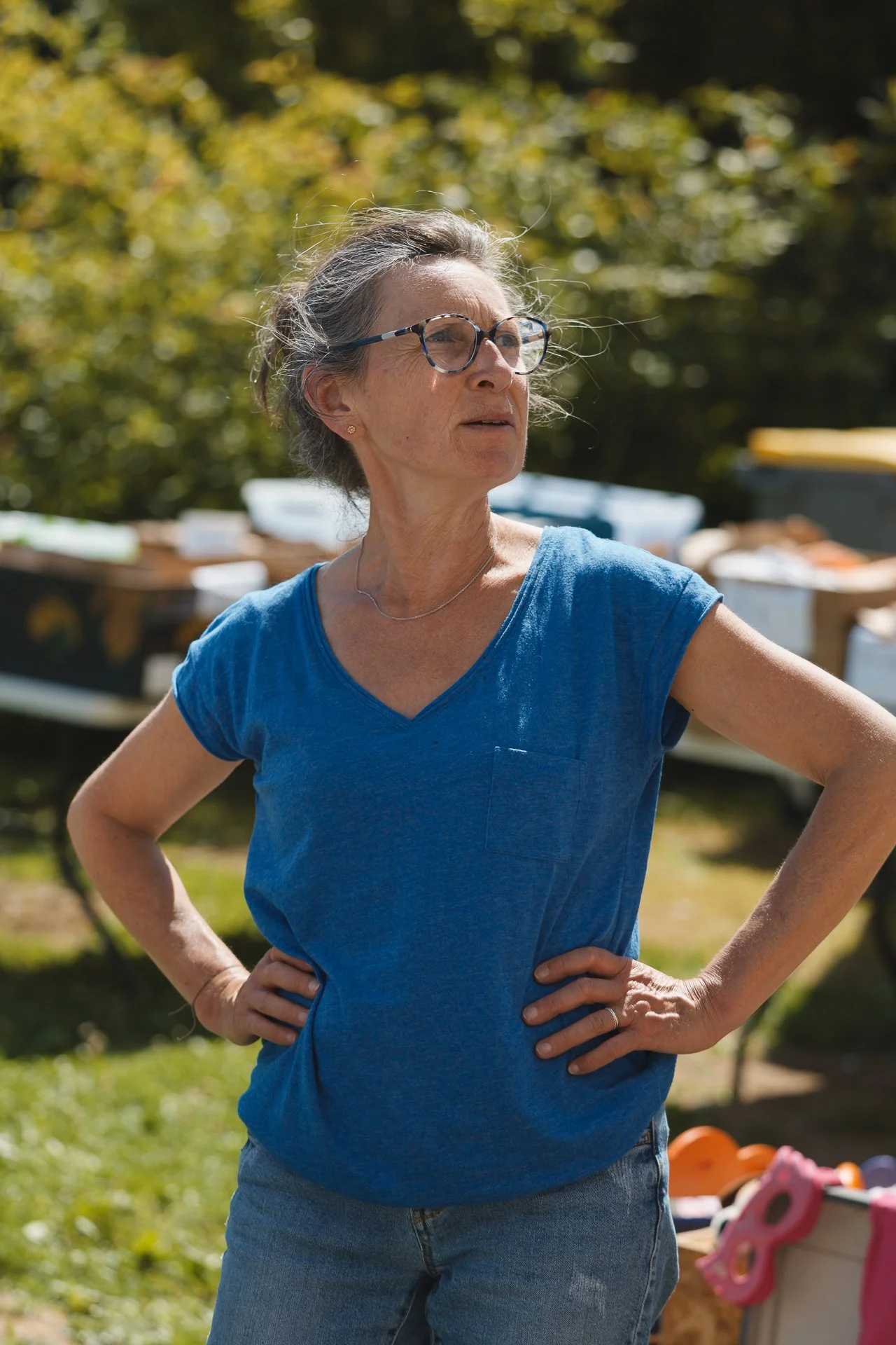Une femme debout en plein air avec ses mains sur ses hanches, portant des lunettes et un t-shirt bleu, en arrière-plan un arbre et des jouets.