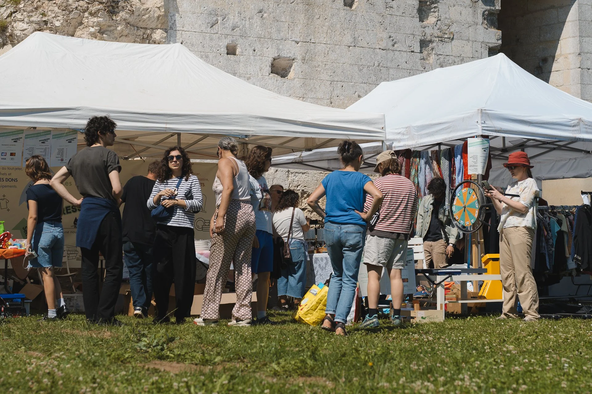 Groupe de personnes faisant du shopping lors d'un marché en plein air, sous des tentes blanches, avec un mur en pierre en arrière-plan.
