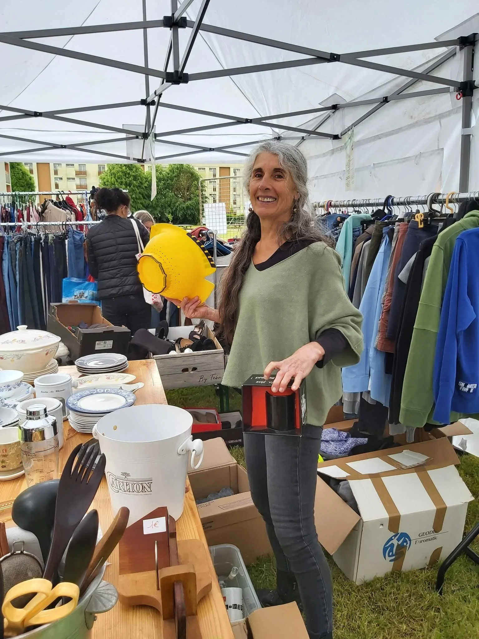 Une femme souriante tenant une tête de casque jaune lors d'un vide-greniers sous une tente blanche, avec des vêtements et des objets divers exposés autour.