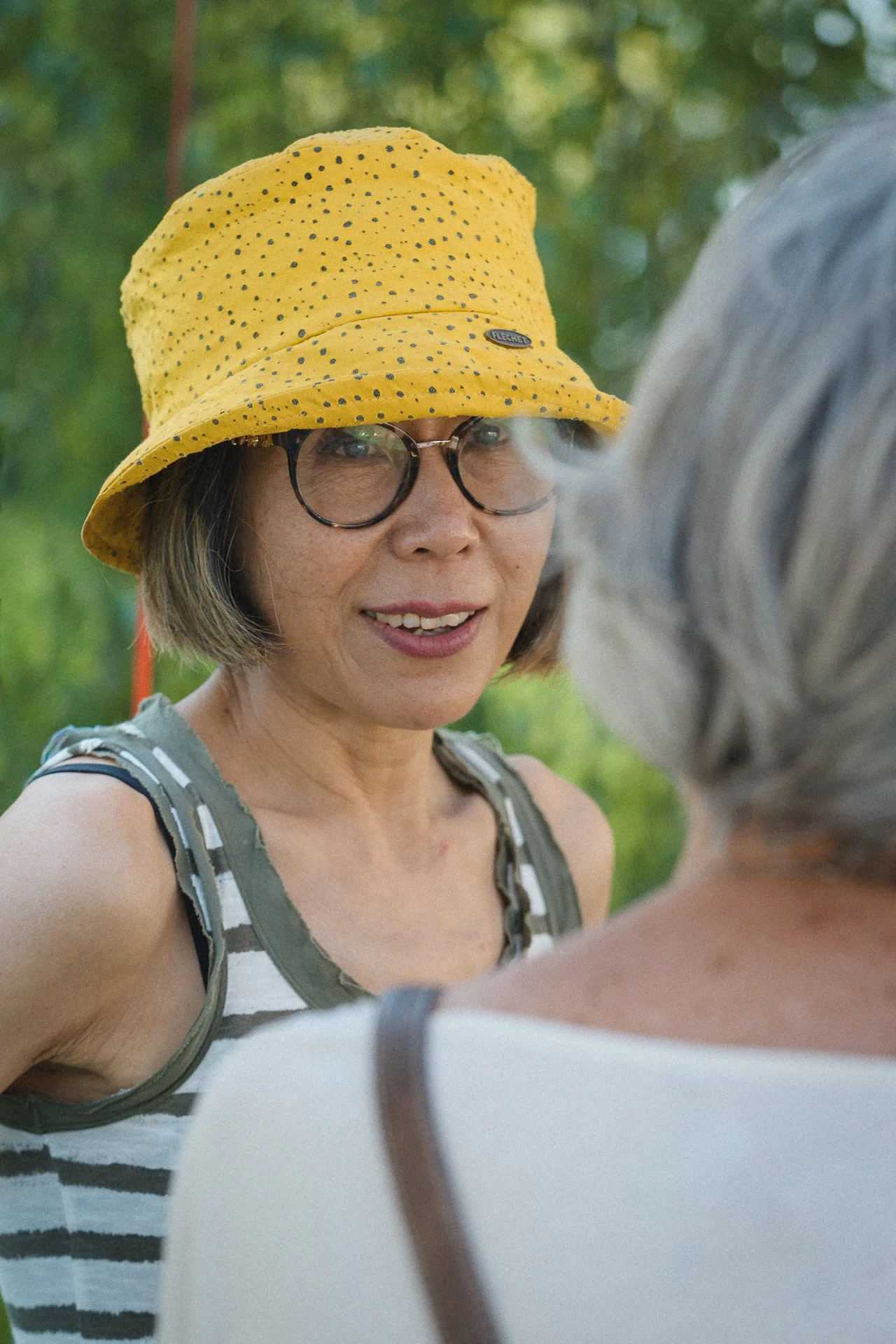 Une femme portant un chapeau jaune à pois noirs, des lunettes rondes, un haut à rayures gris et blanc, et souriant, elle est en conversation avec une autre personne en arrière-plan.