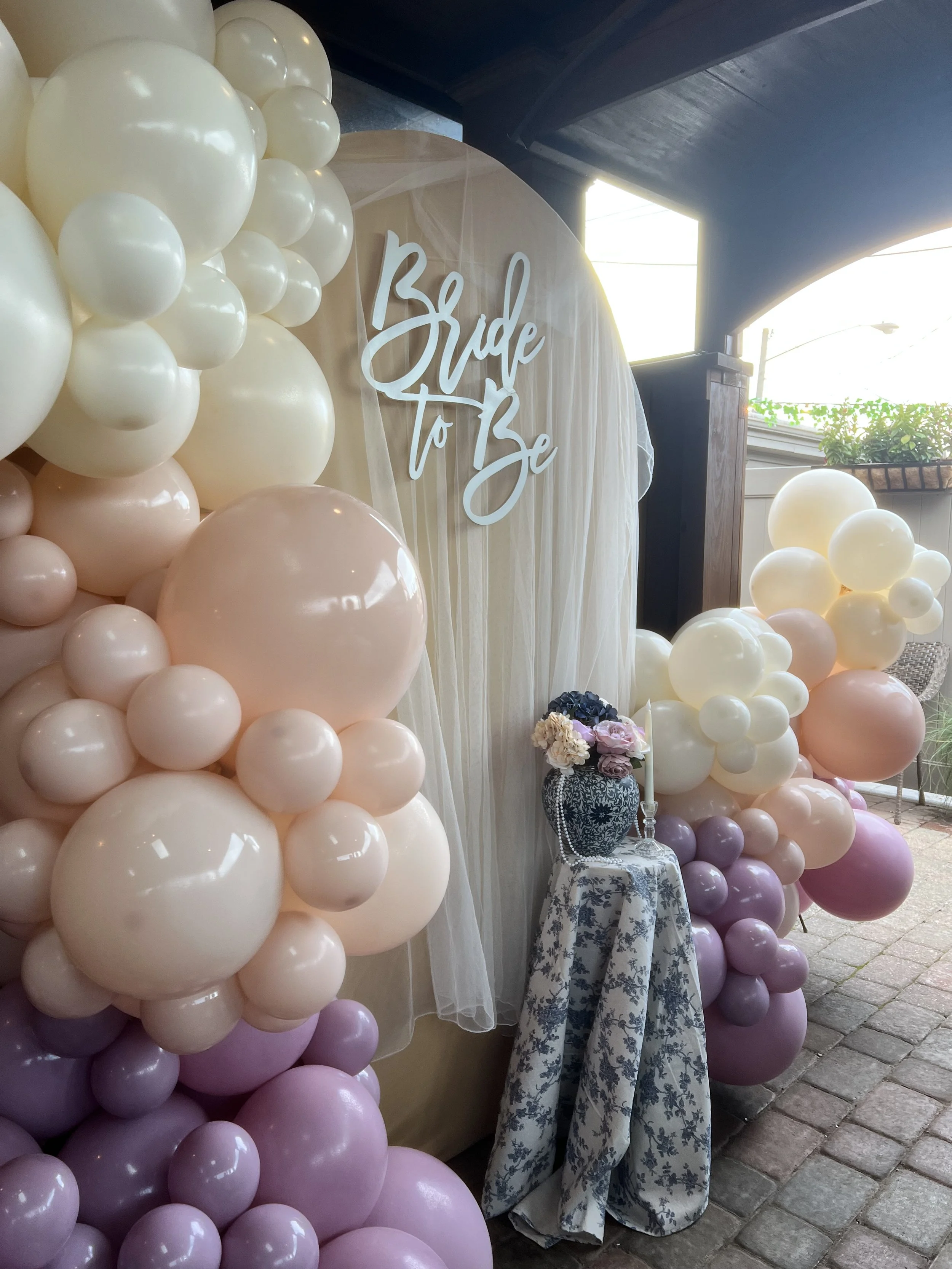 Decorative setup with pink and white balloons, a sign that reads 'Bride to Be,' a table with flowers and a candle, outdoor setting with a cobblestone ground.