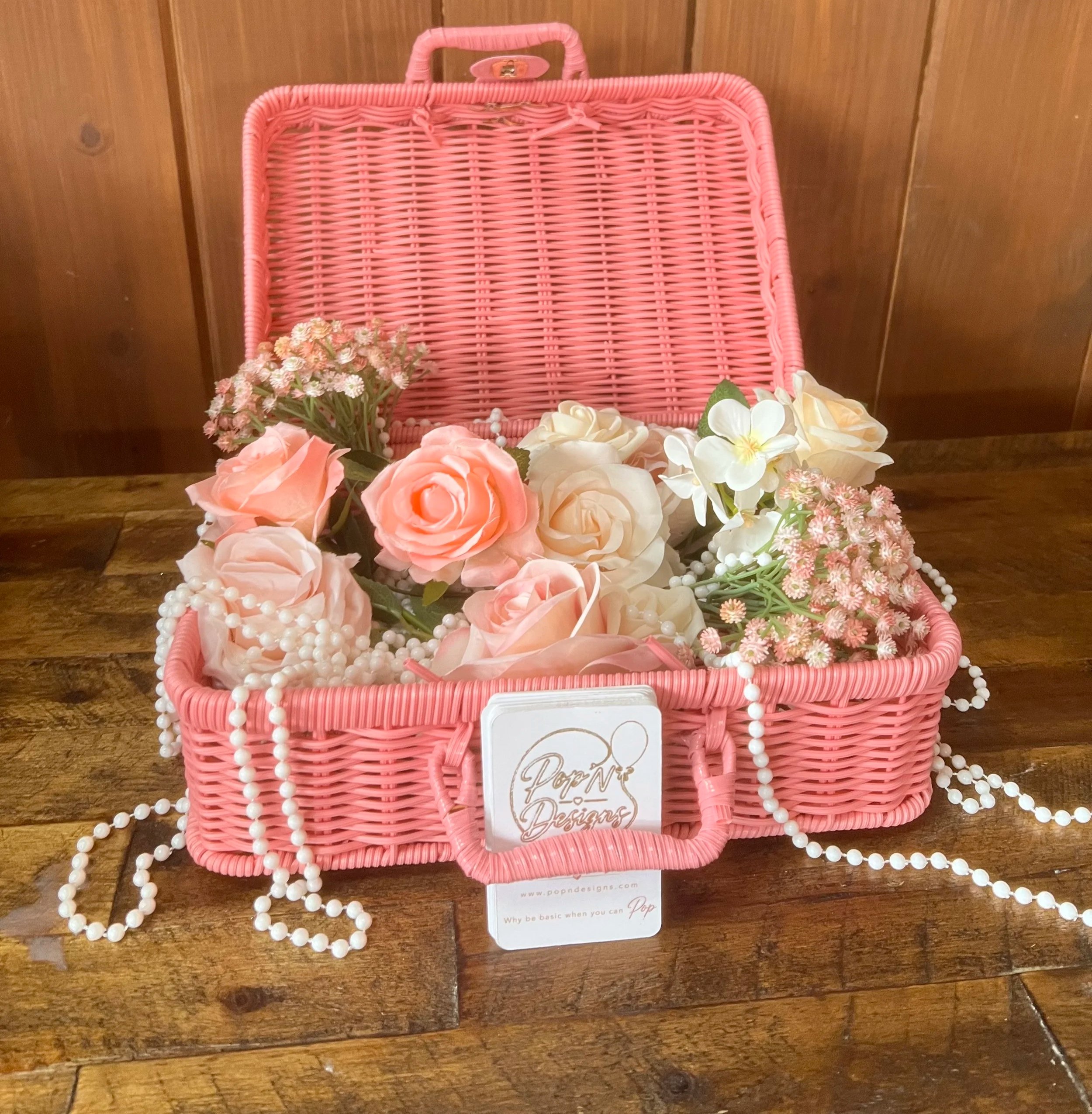 A pink woven basket filled with pink and white artificial flowers, jewelry, and a business card, placed on a wooden surface against a wooden wall background.