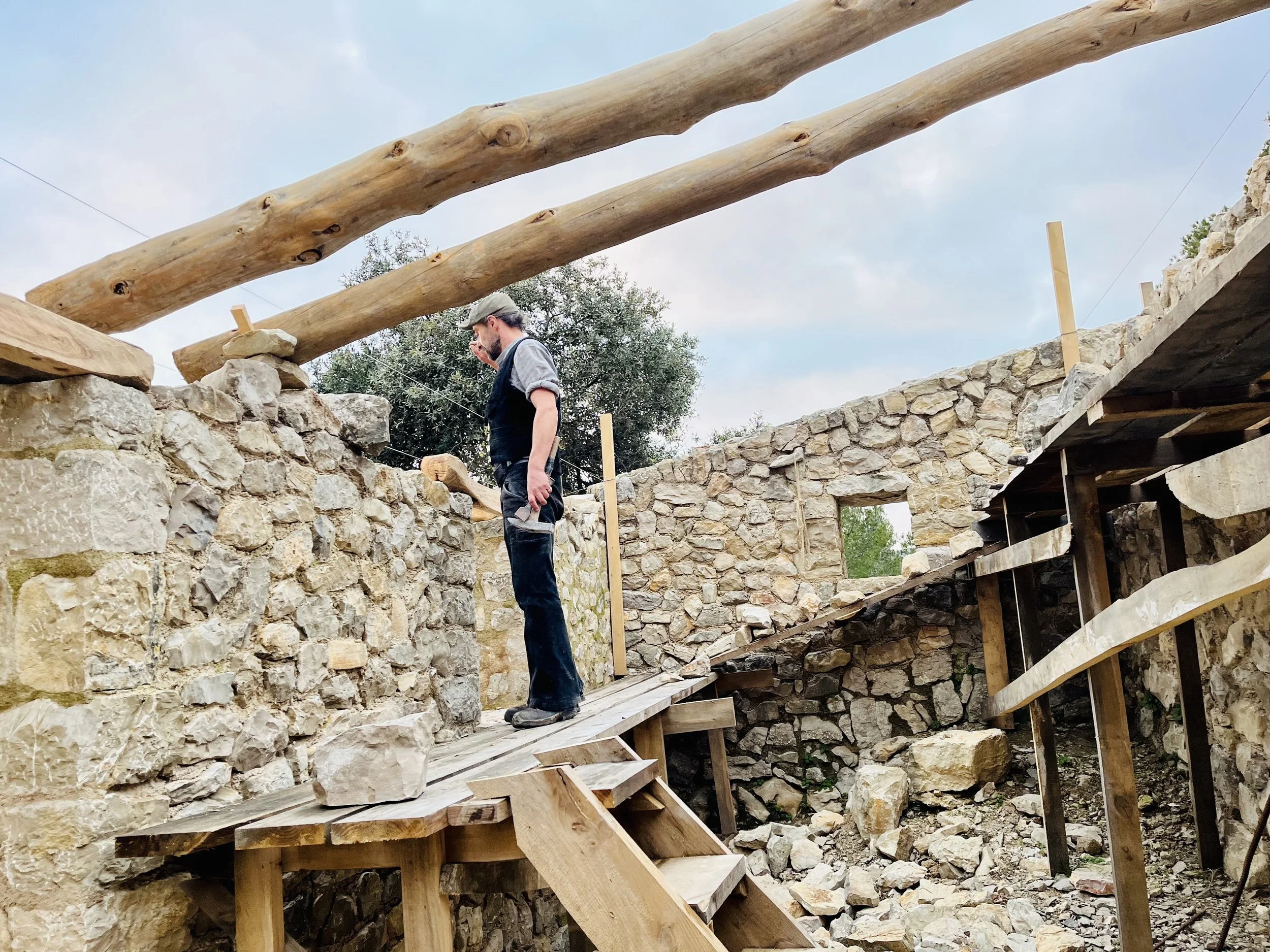 A man wearing a hat, gray shirt, and dark overalls stands on a wooden platform overseeing stonework construction, with large wooden beams positioned overhead and stone walls surrounding him.
