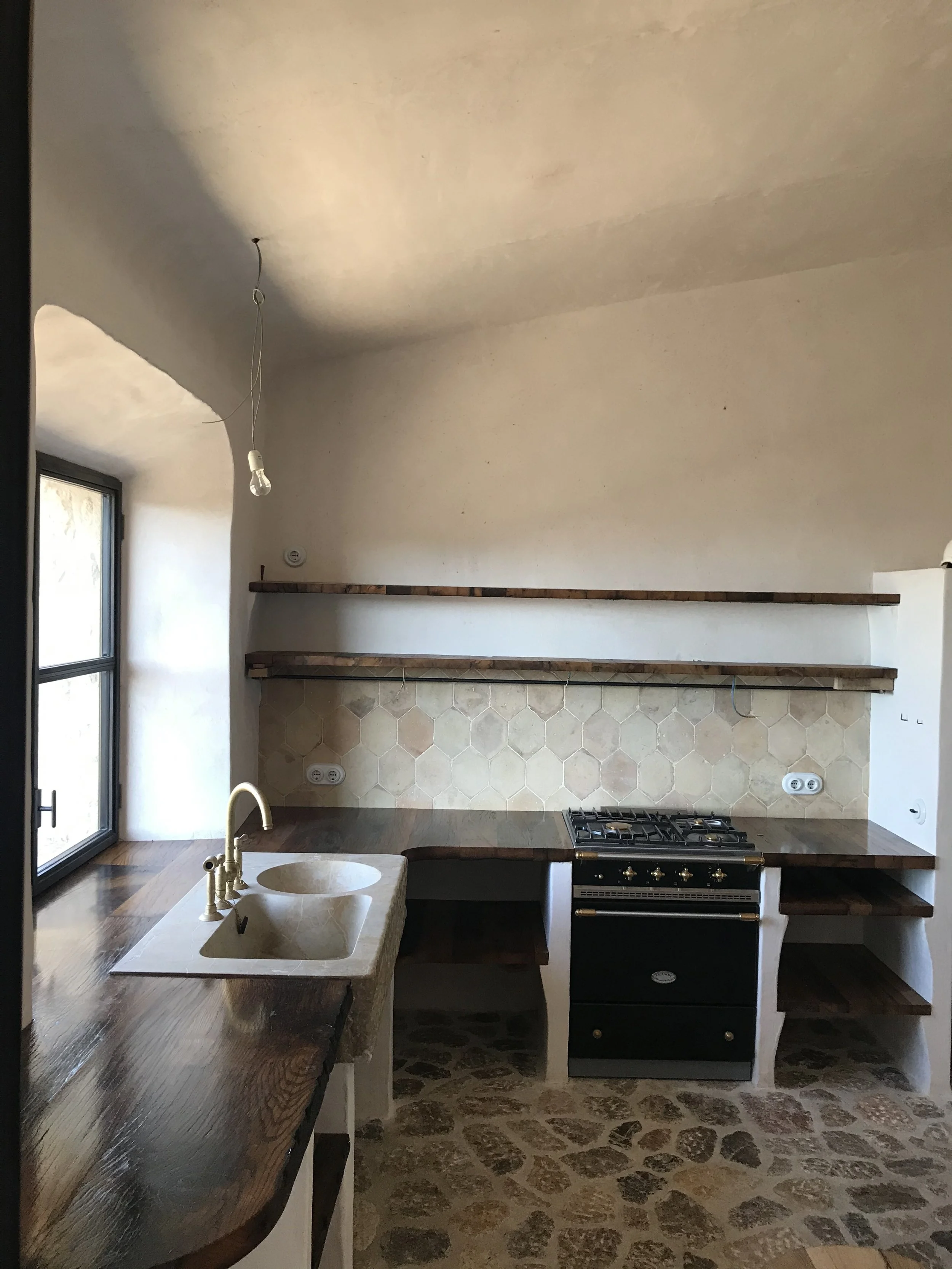 A kitchen with a wooden countertop, a built-in sink with a brass faucet, a gas stove with gold knobs, a stone tile backsplash, and a cobblestone floor. There are shelves on the wall above the counter, electrical outlets, and a window to the left.