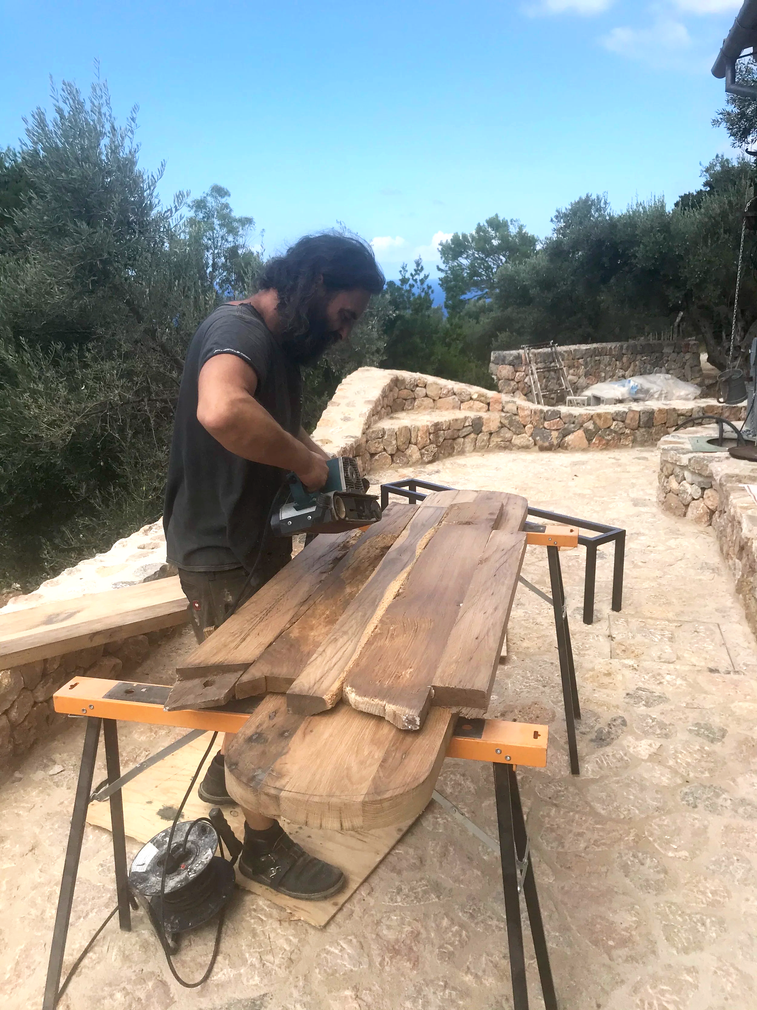A man working outdoors on woodworking projects, cutting large wooden planks on a workbench, with stone structures and trees in the background.