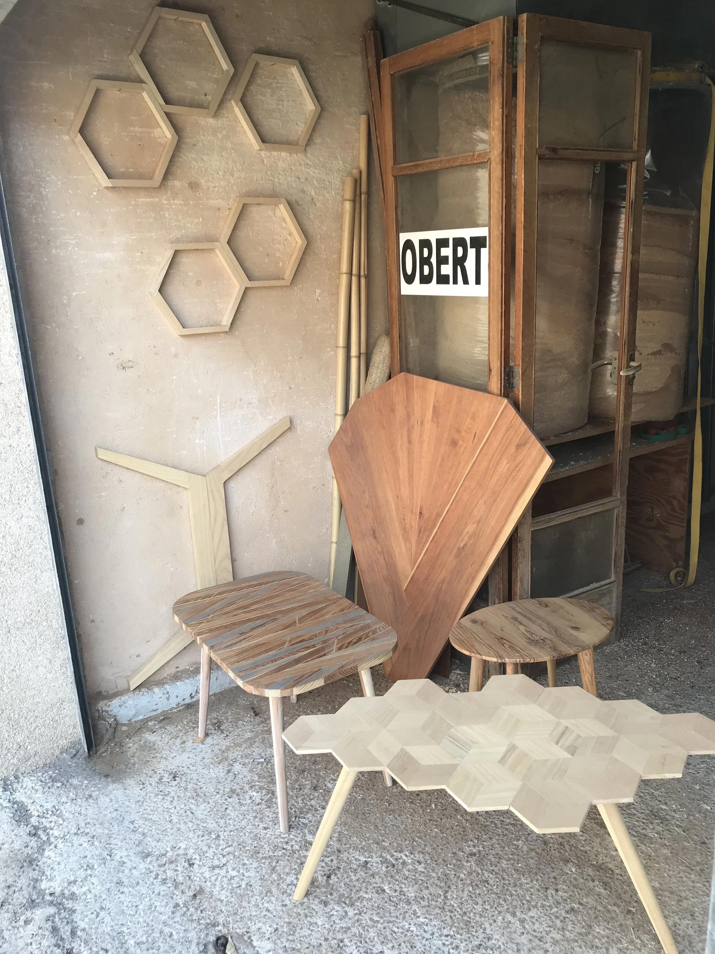 Various wooden furniture pieces including hexagonal tables and chairs are displayed, with some items on the ground and others on a shelf, in a workshop.