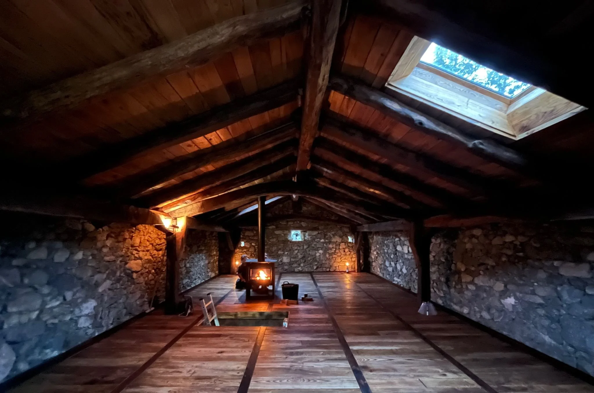 Wooden ceiling with large beams and a skylight letting in natural light. Stone walls with a camping stove and chairs in a rustic cabin interior.