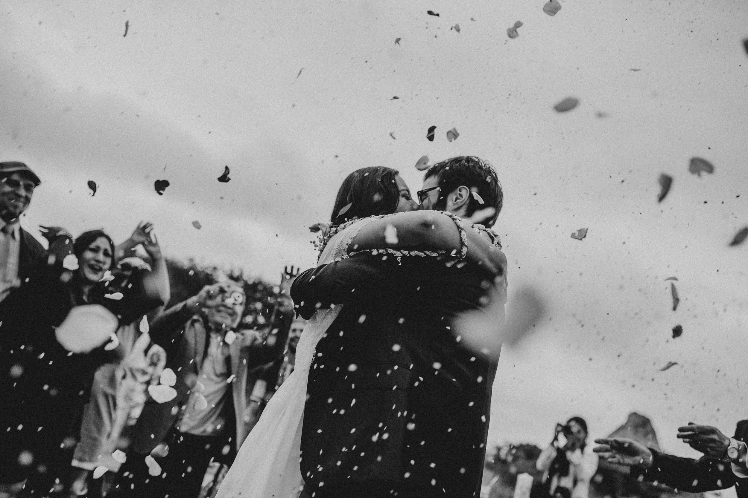 Newly married couple sharing a kiss while the family celebrates and confetti falls from the sky.