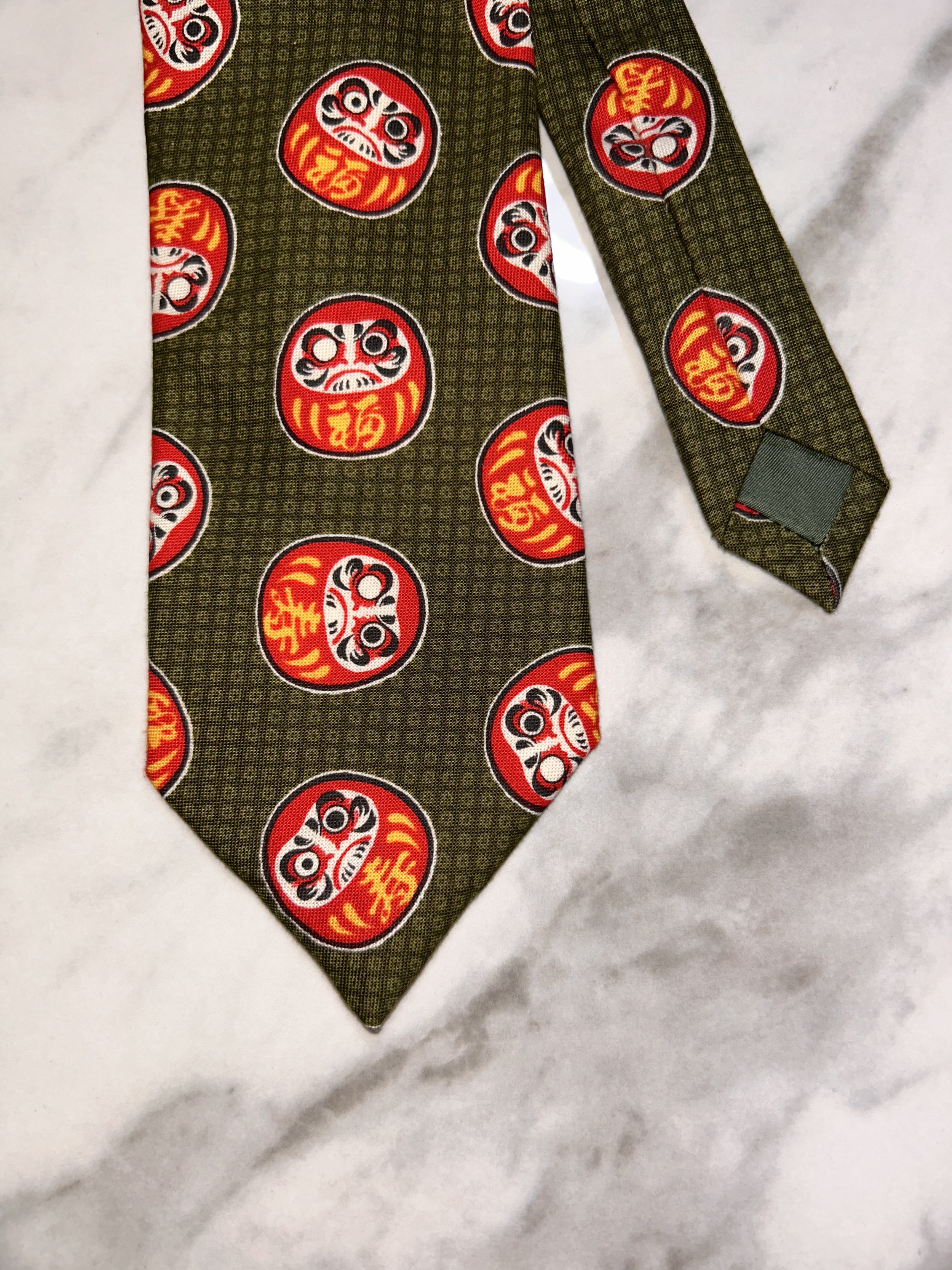 A piece of fabric with a pattern of red and white Daruma dolls on a dark green background, with the fabric folded at the corner.