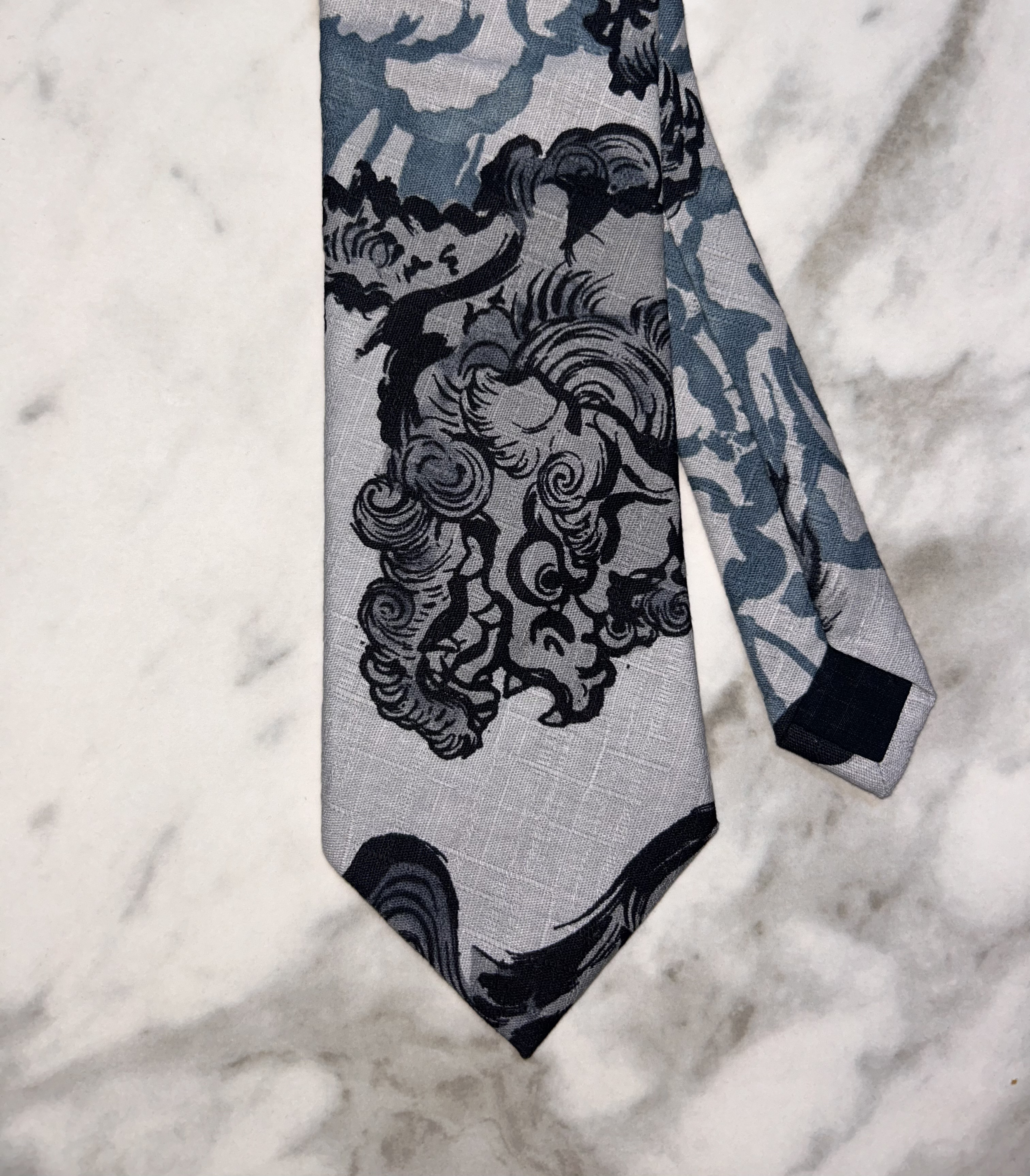 Close-up of a fabric with a detailed black and gray floral or botanical pattern on a light background, with the fabric's edge visible at the bottom of the image.