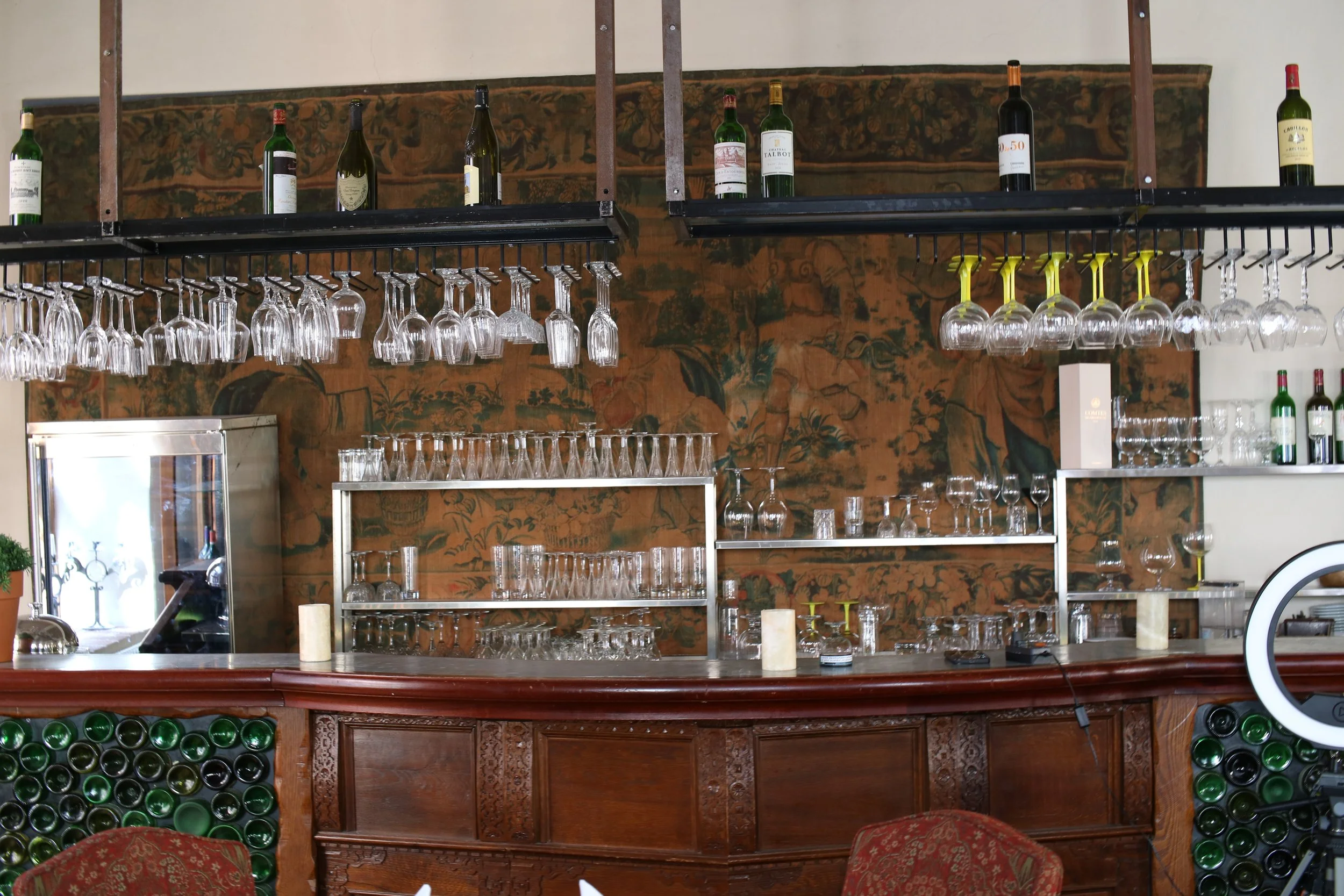 A bar with shelves holding wine bottles and glassware, with a decorative tapestry and bottles stored on the side.