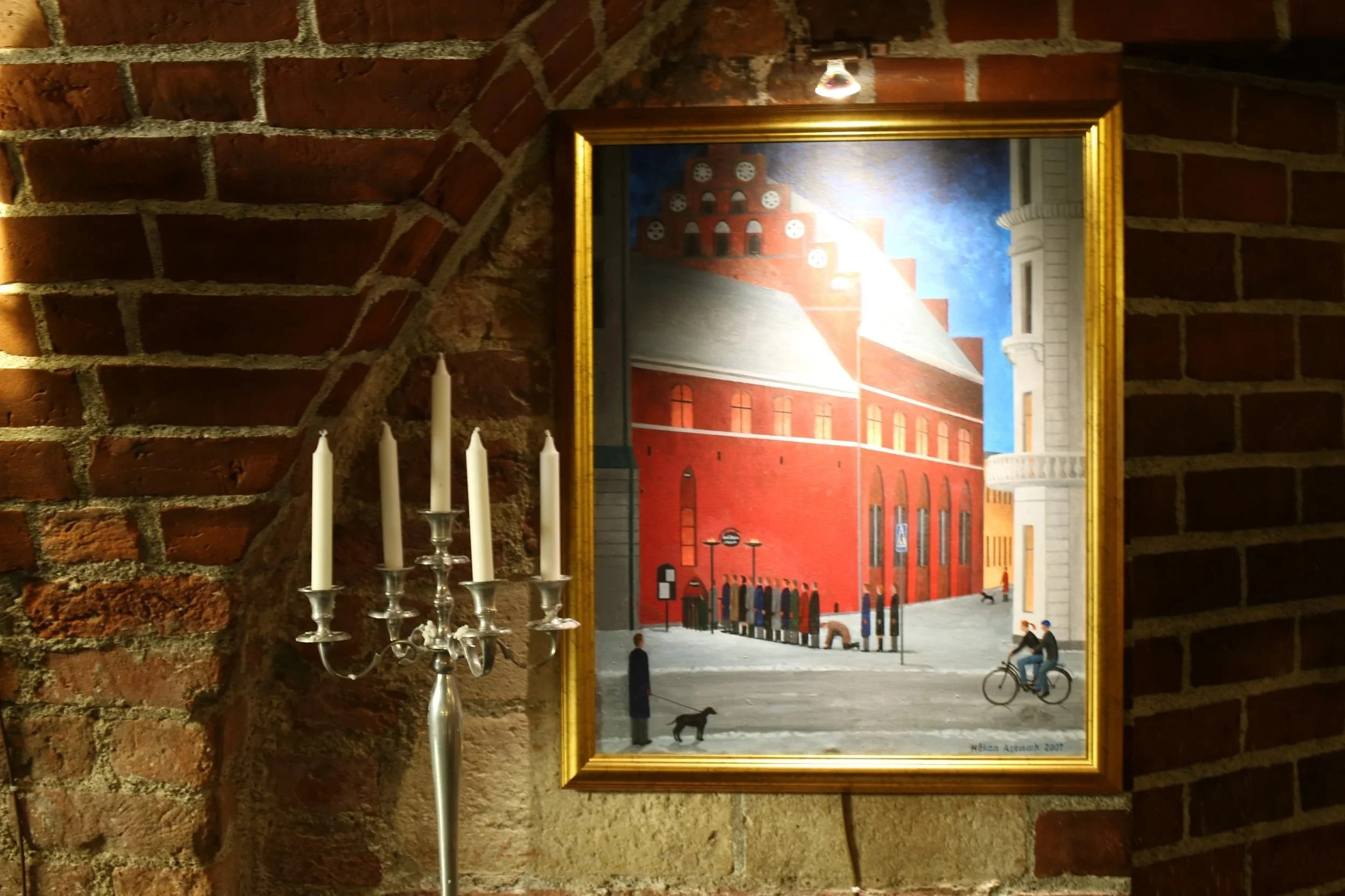 A painting is hanging on a brick wall showing a cityscape with red brick buildings and people walking. A black dog and a person on a bicycle are visible in the scene.