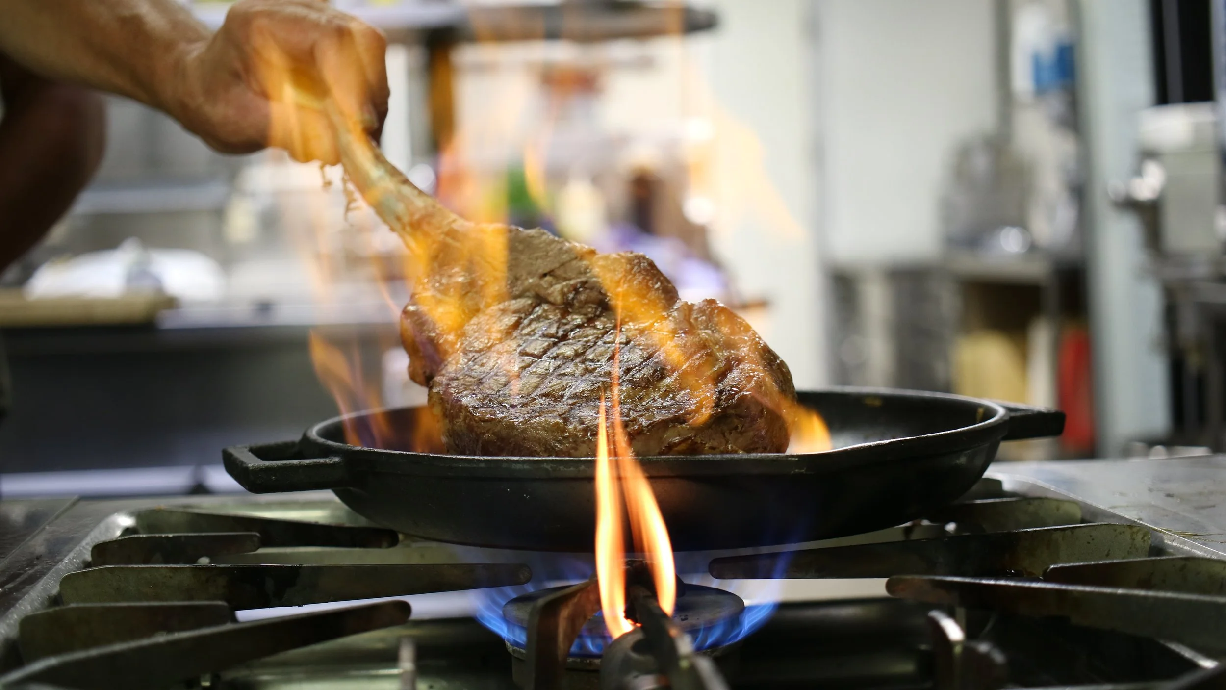 A piece of steak being cooked on a stovetop grill with flames, with a hand holding a wooden spatula or fork nearby.