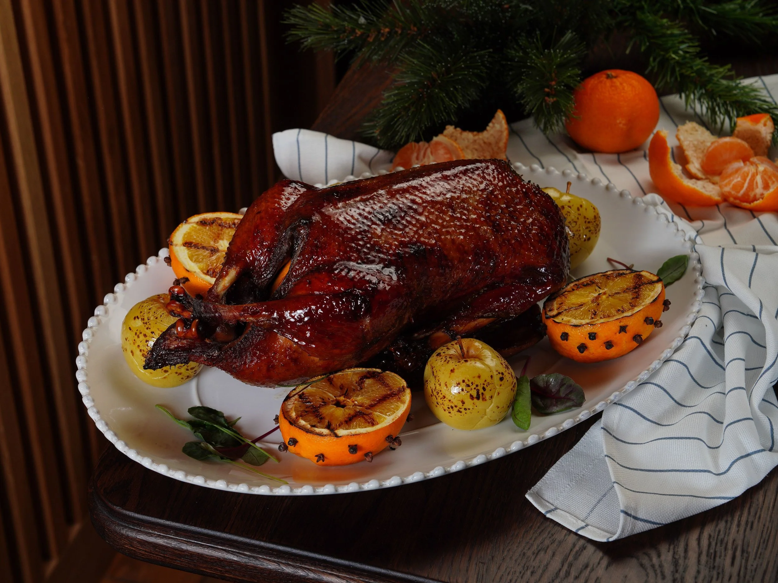 A roasted turkey garnished with grilled orange slices, surrounded by tangerines and greenery on a white decorative platter, with a blue and white striped cloth nearby.