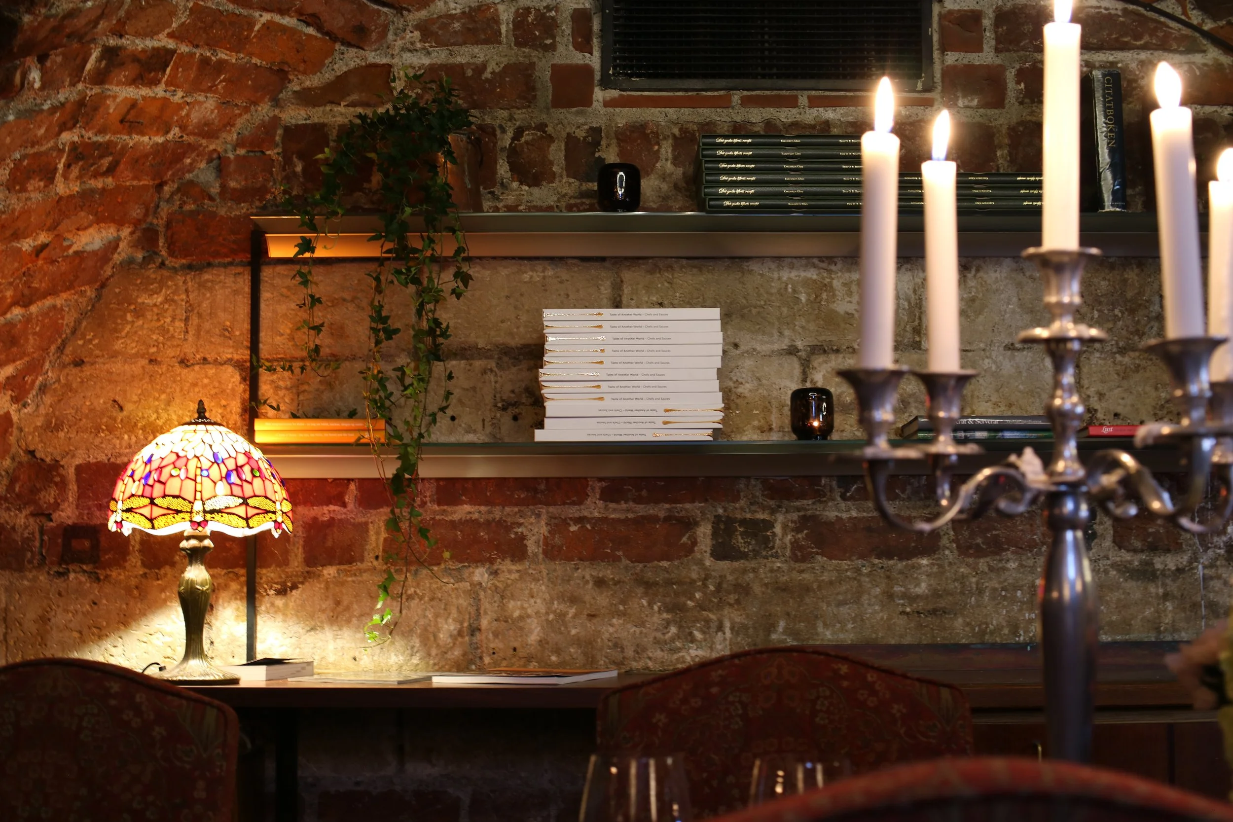 A cozy interior scene with a brick wall, a stained glass table lamp, a shelf with books and plants, and several lit candles on a chandelier in the foreground.