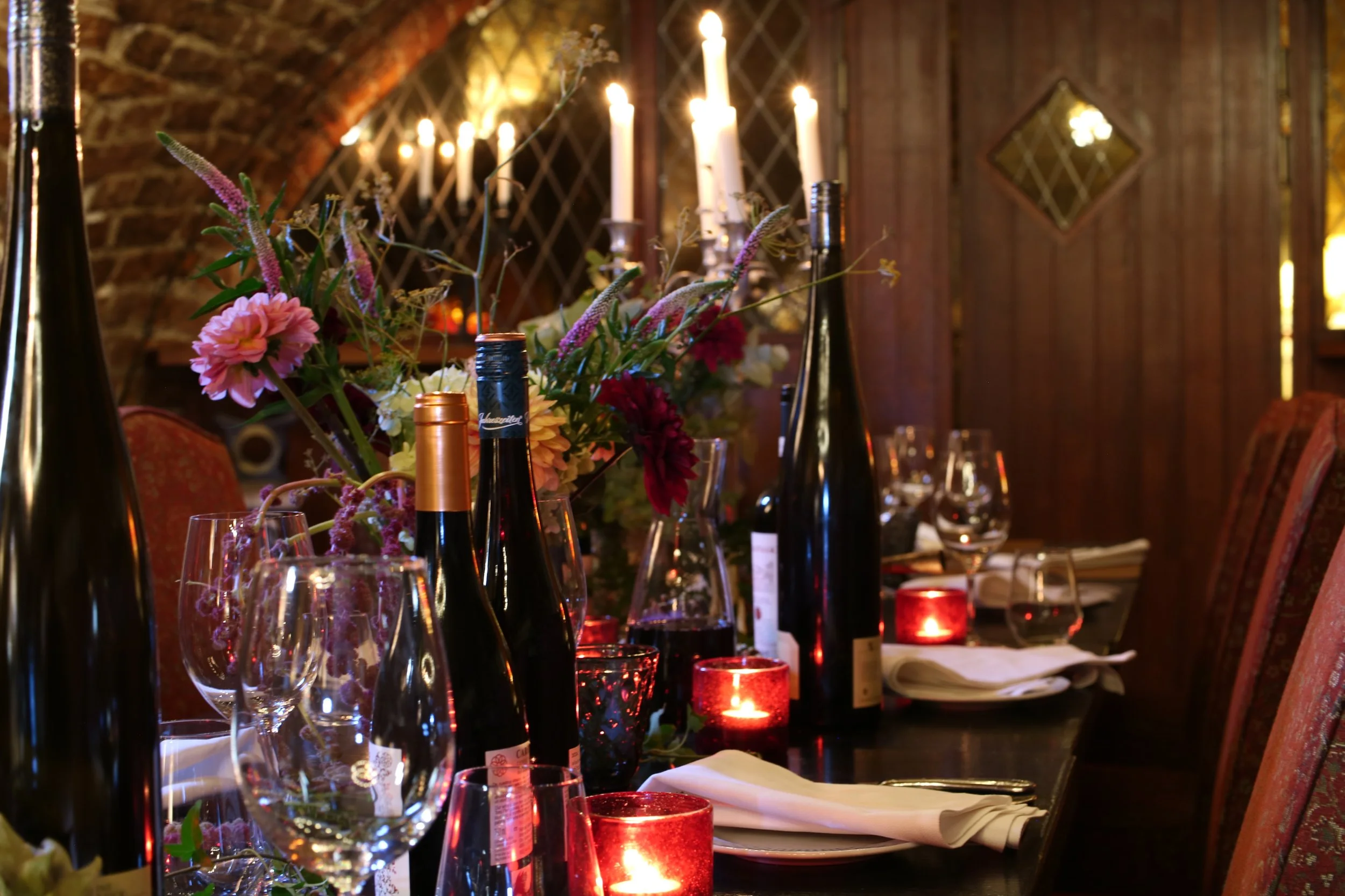 A warmly lit dining table decorated with a floral centerpiece, wine bottles, wine glasses, candles, and tableware in a rustic brick-walled room.