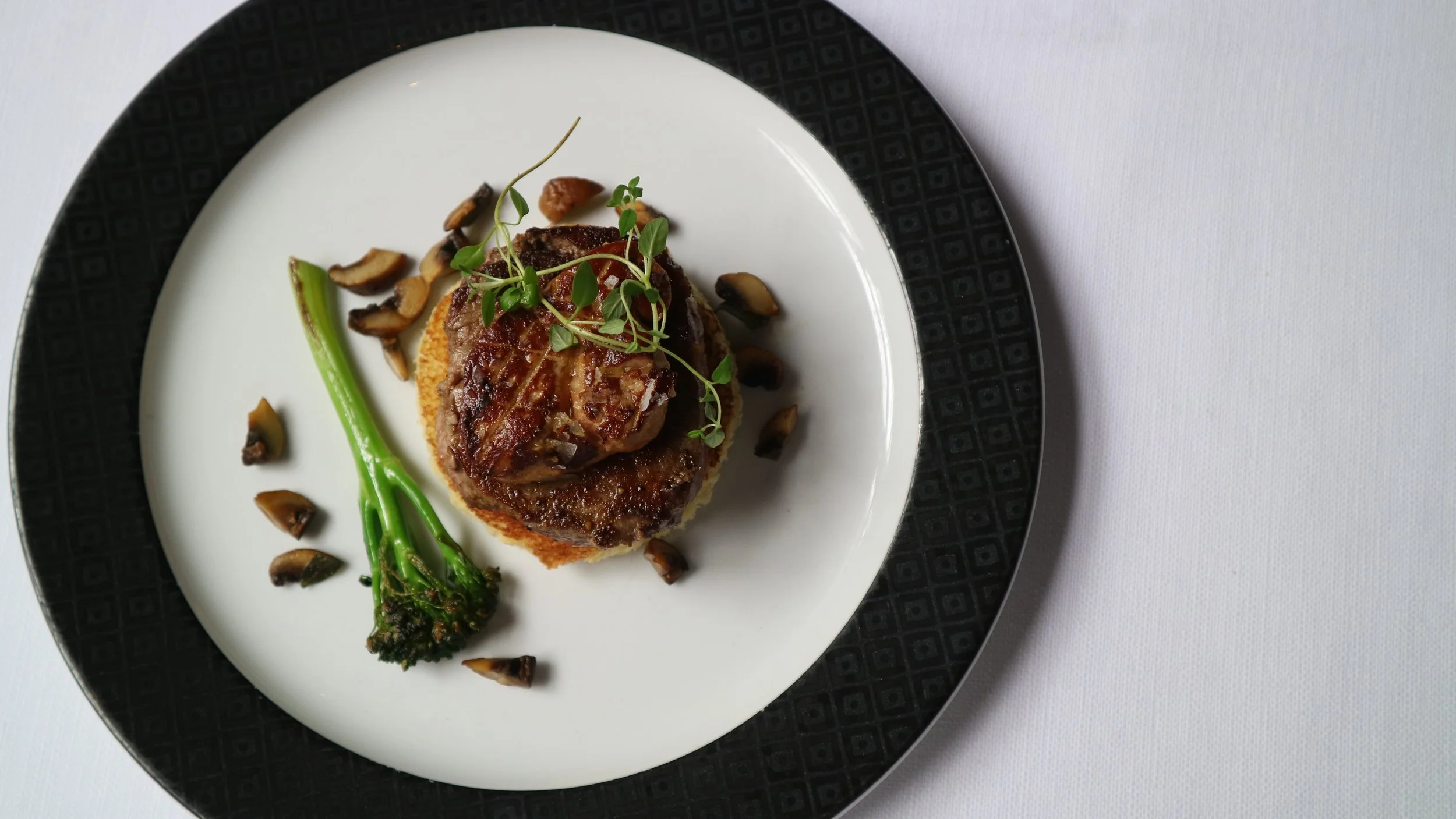 A plated gourmet dish featuring a piece of meat topped with microgreens, served over a bed of mashed potatoes or polenta, garnished with roasted or sautéed mushrooms, green beans, and a small piece of roasted broccoli, on a black and white patterned plate.