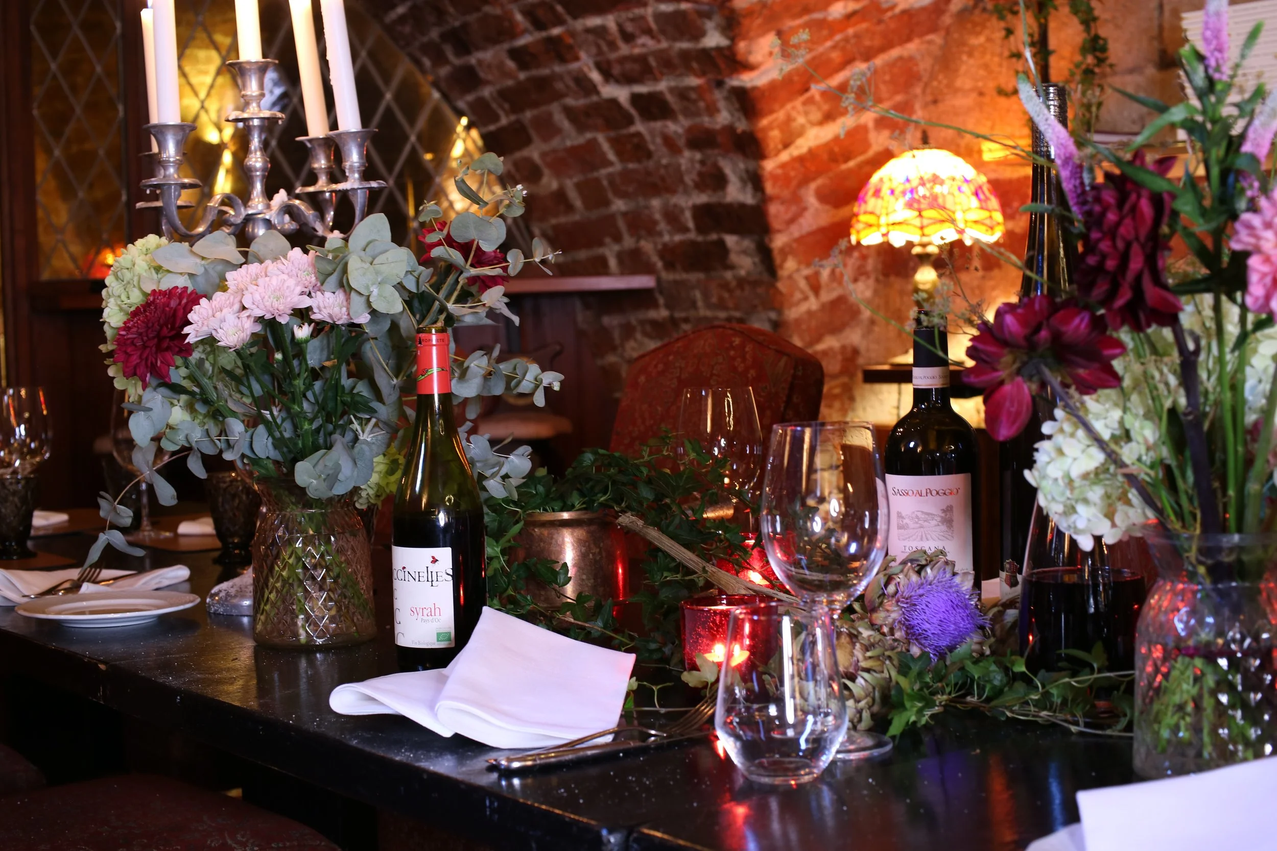 A decorated dining table with floral arrangements, wine bottles, glassware, and lit candles, set against a brick wall with warm lighting.