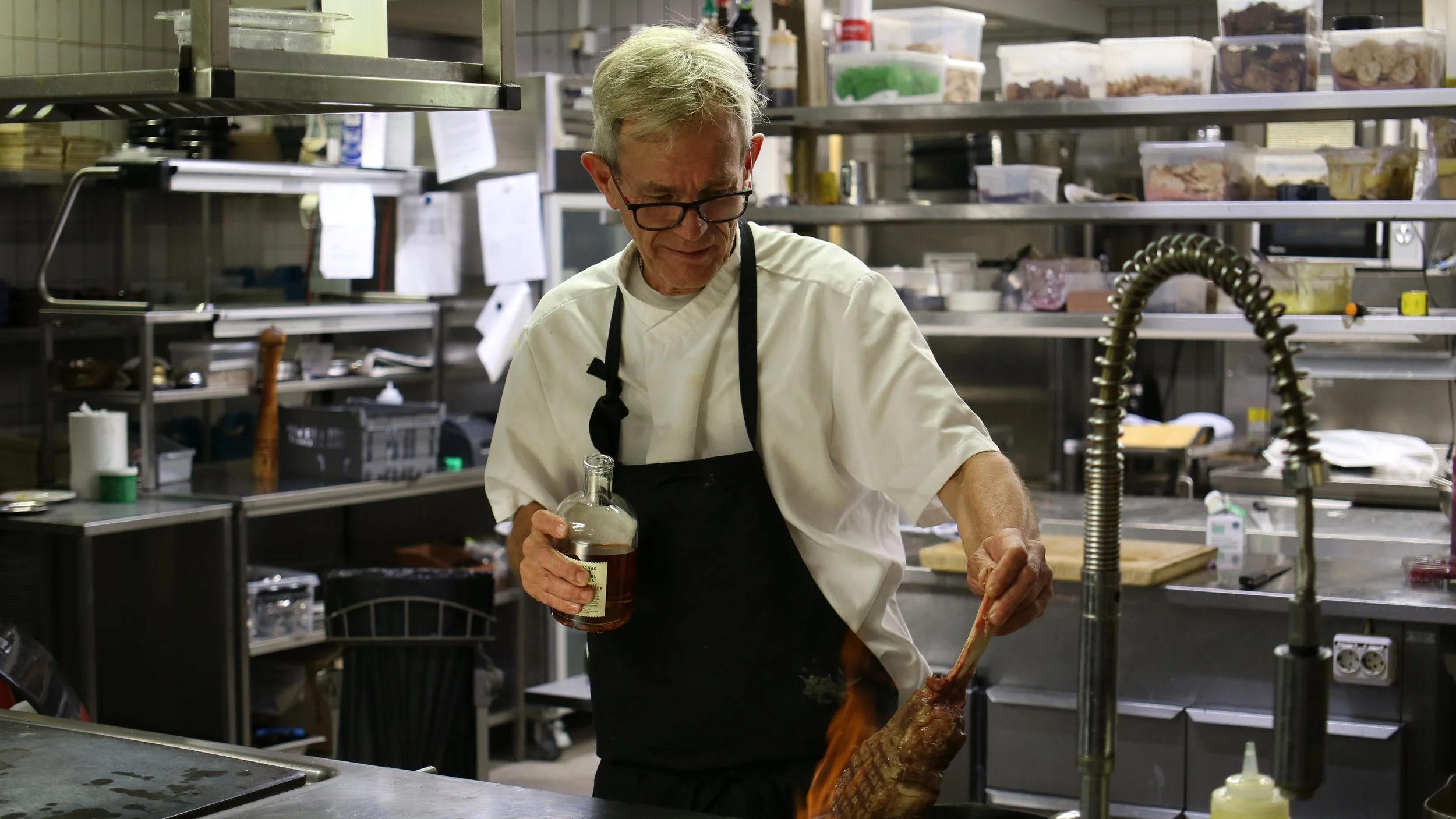 An elderly man with gray hair, glasses, wearing a white chef's coat and black apron, standing in a commercial kitchen, holding a bottle of liquid and a skewer with meat, preparing food.