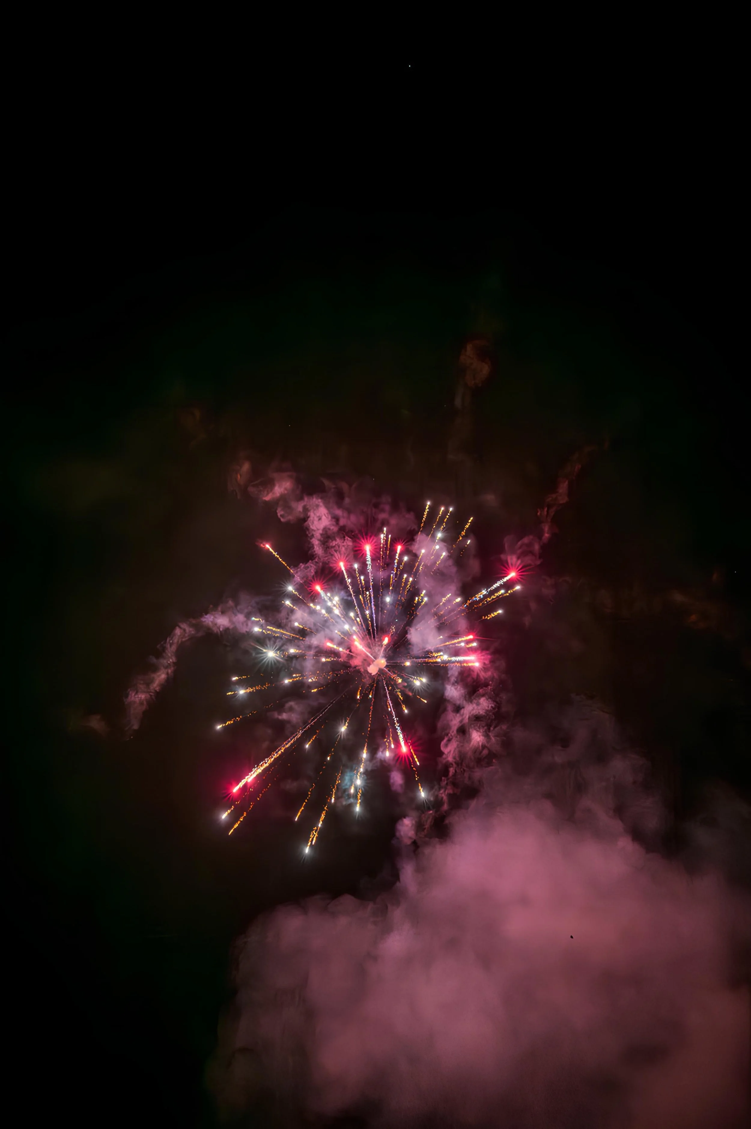 Colorful fireworks exploding in the night sky with pink, white, and yellow streaks amid clouds of smoke.